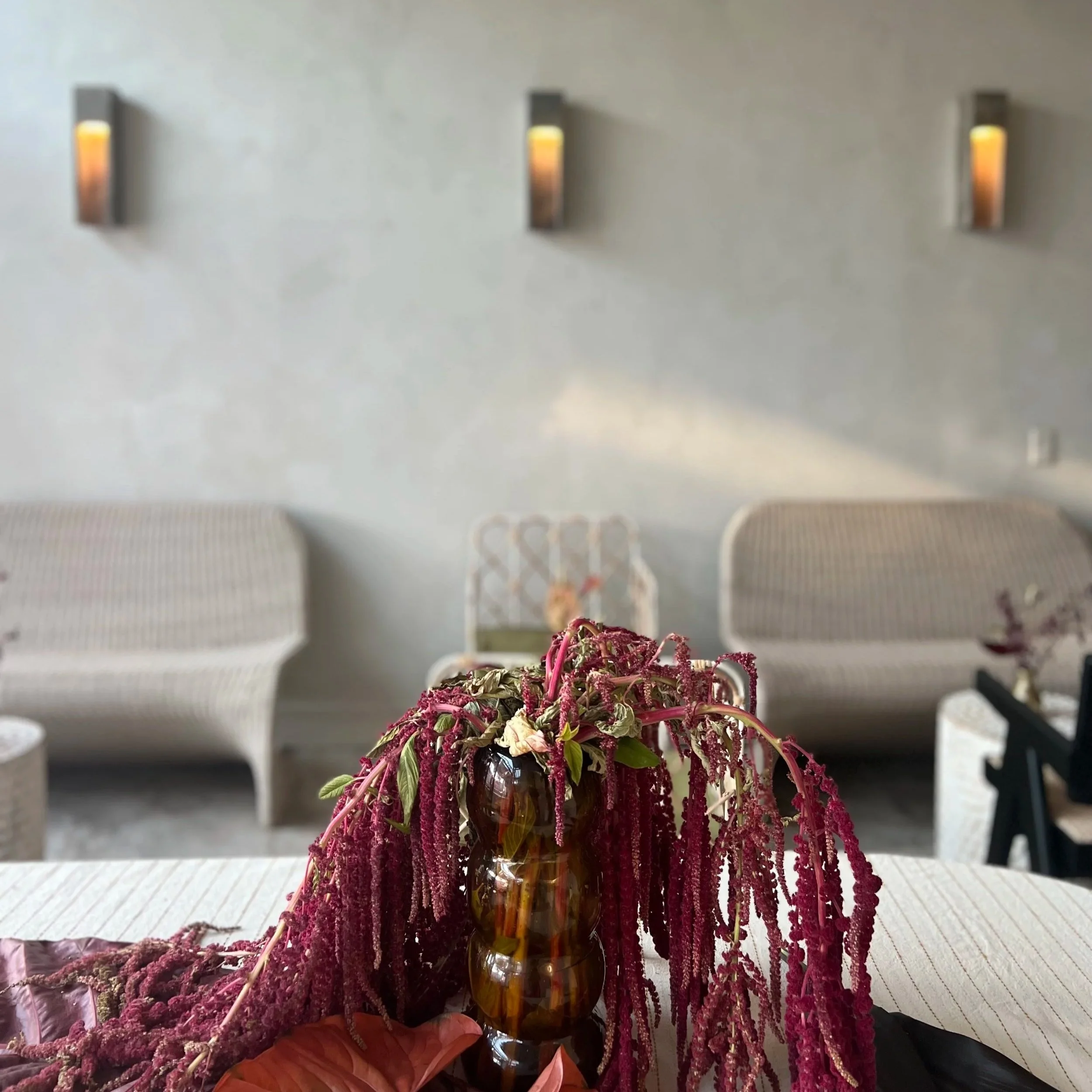 A floral arrangement in a glass vase with purple and green flowers and hanging purple amaranthus, placed on a table in a room with upholstered chairs and wall-mounted lights in the background.