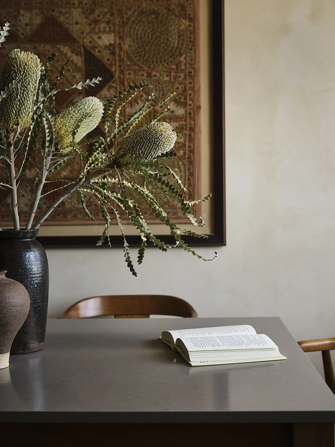 A minimalist table with an open book, a vase with green leaves and protea flowers, set against a wall with a framed textile artwork.