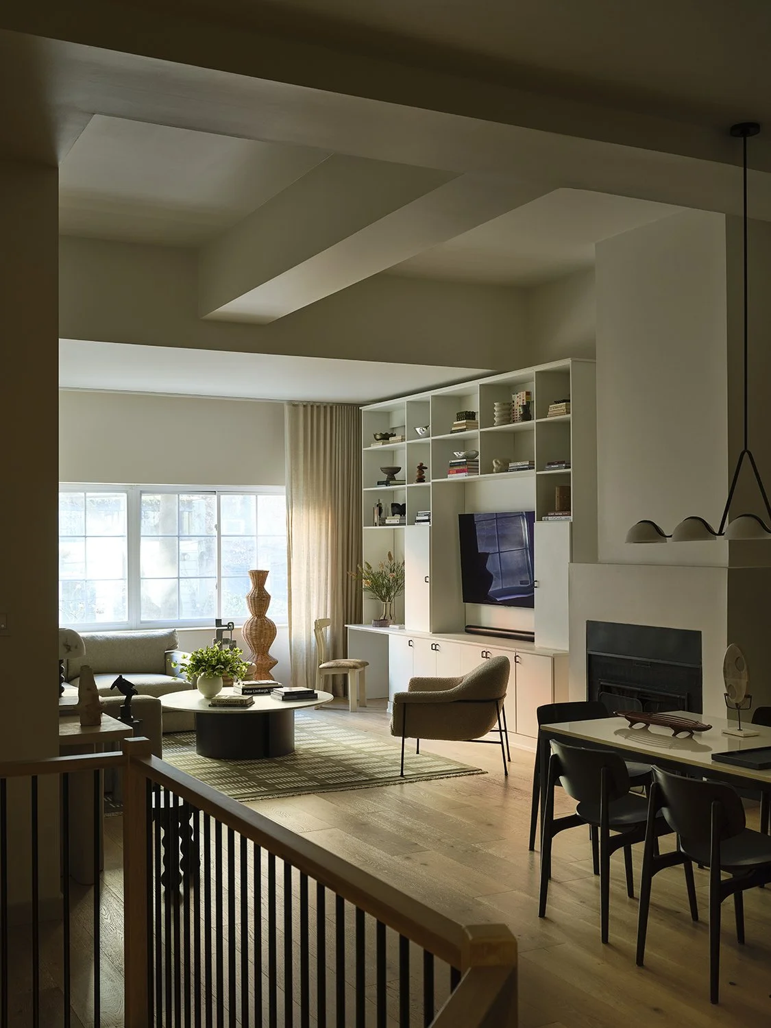 A cozy living room with large window, beige curtains, white shelving unit, a TV, armchair, coffee table, and decorative items.