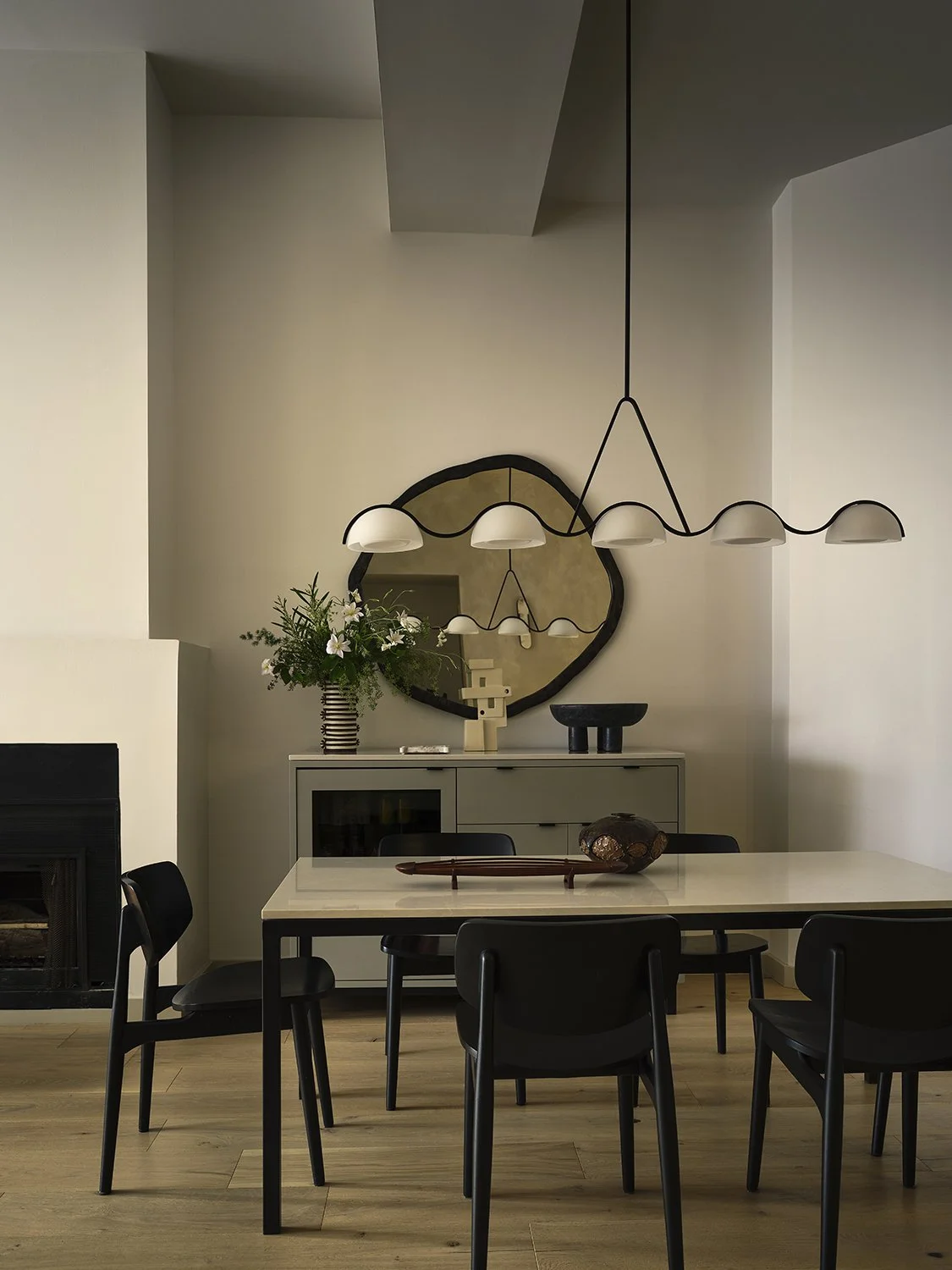 Modern dining room with a rectangular table, six black chairs, a sideboard with decorative objects, a plant arrangement, a mirror, and a sculptural chandelier hanging from the ceiling.