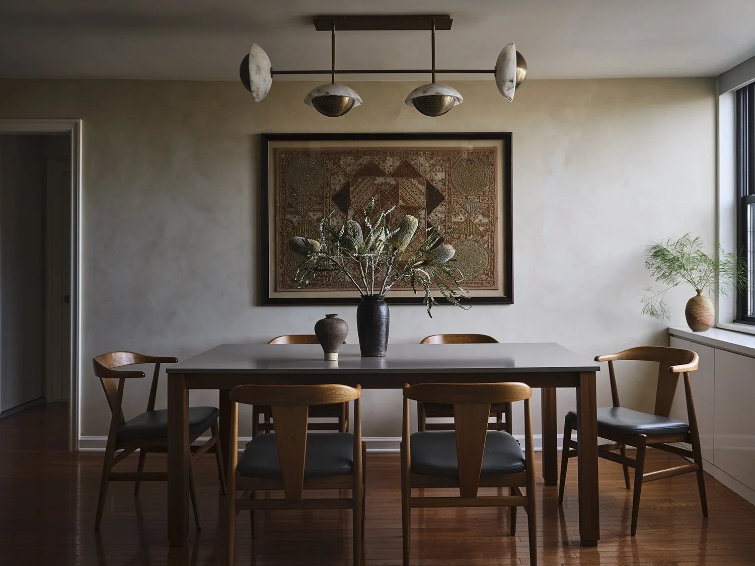 A dining room with a white table, six wooden chairs, a black vase with dried flowers on top, and framed artwork on the wall. There is a window with a plant on the windowsill.