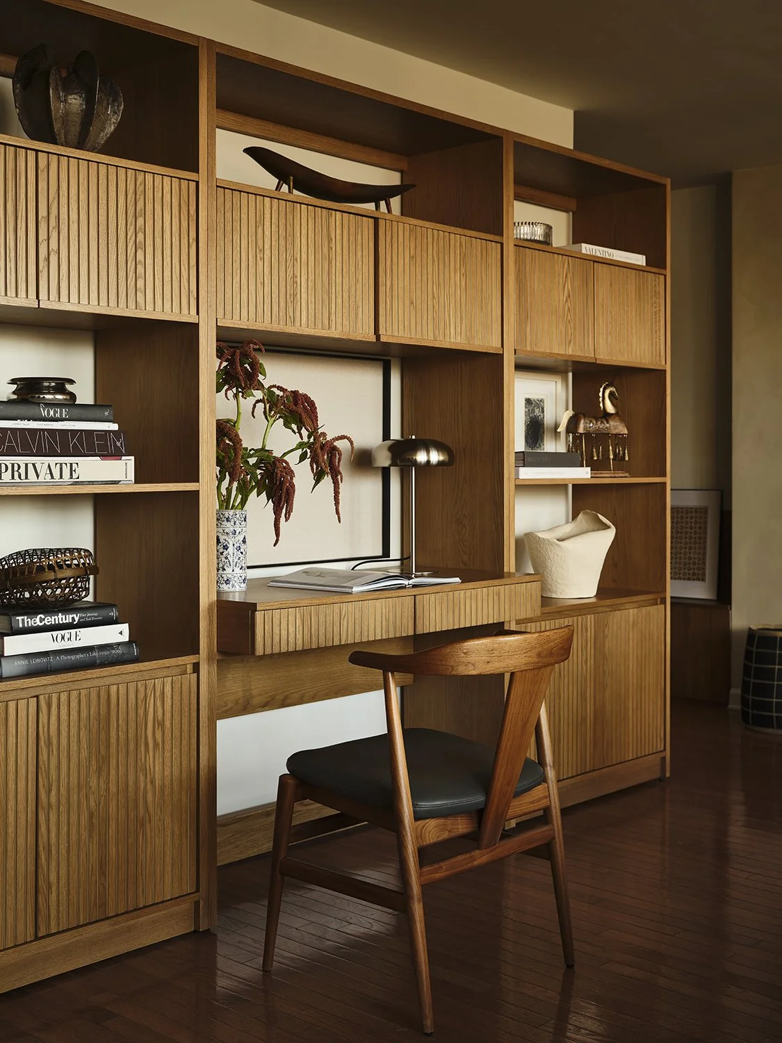 A wooden desk with a black chair in front of a built-in wooden shelving unit decorated with books and decorative objects.