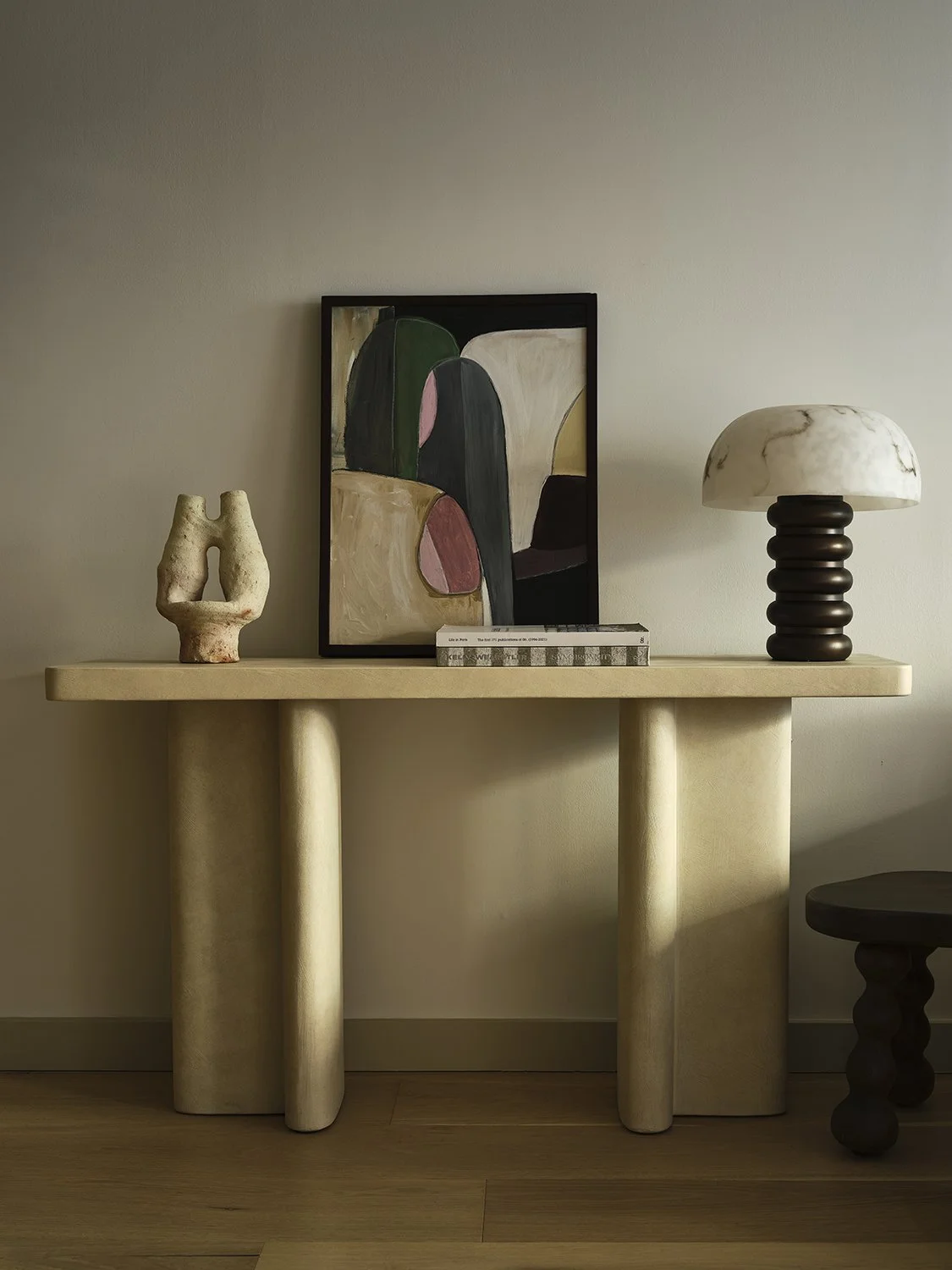 Decorative console table with a framed abstract painting, a textured sculpture, a stack of books, and a table lamp with a cream-colored shade and dark base.