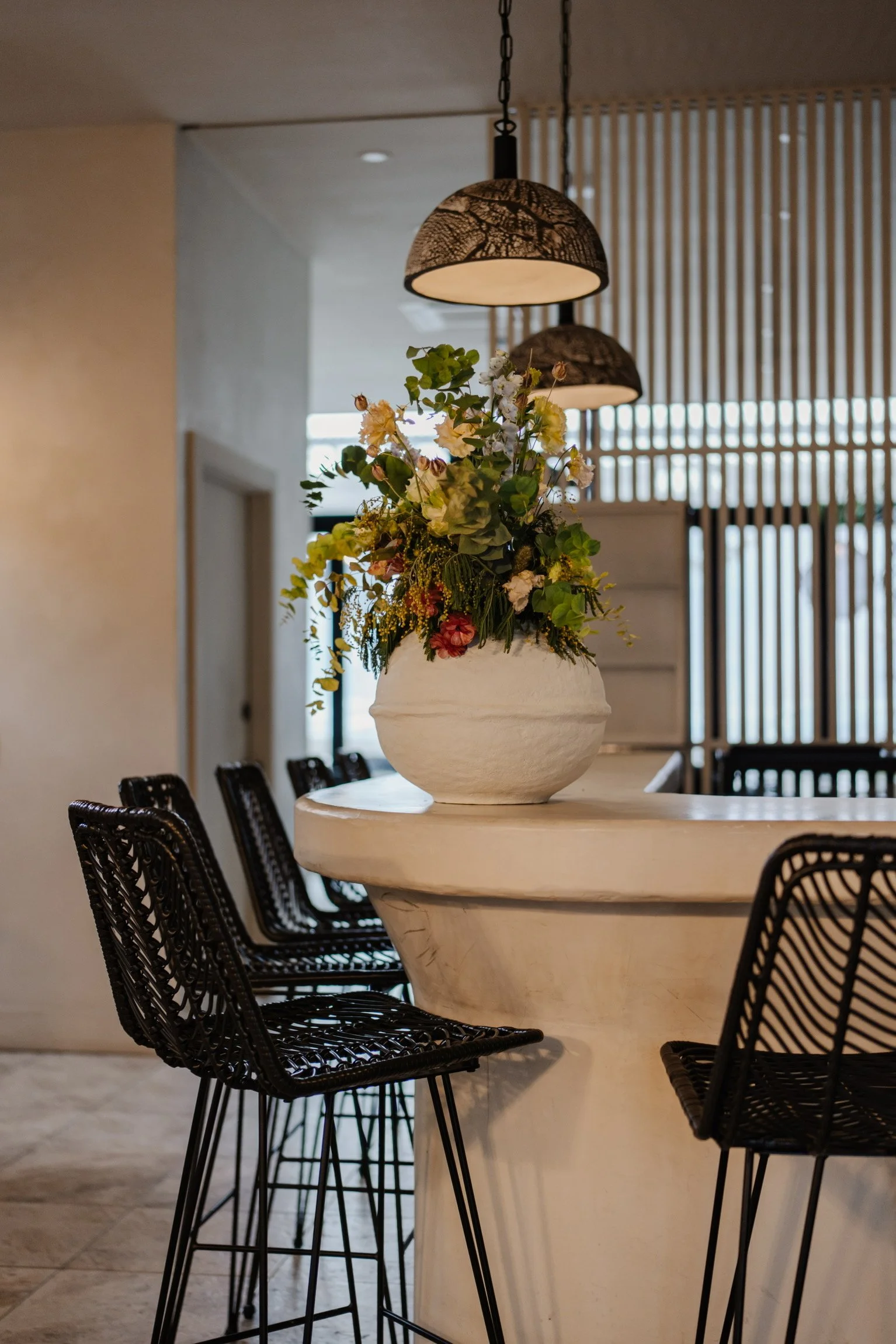 A white round table with black wicker chairs surrounding it, decorated with a large white ceramic vase filled with colorful flowers in an indoor setting with hanging pendant lights.