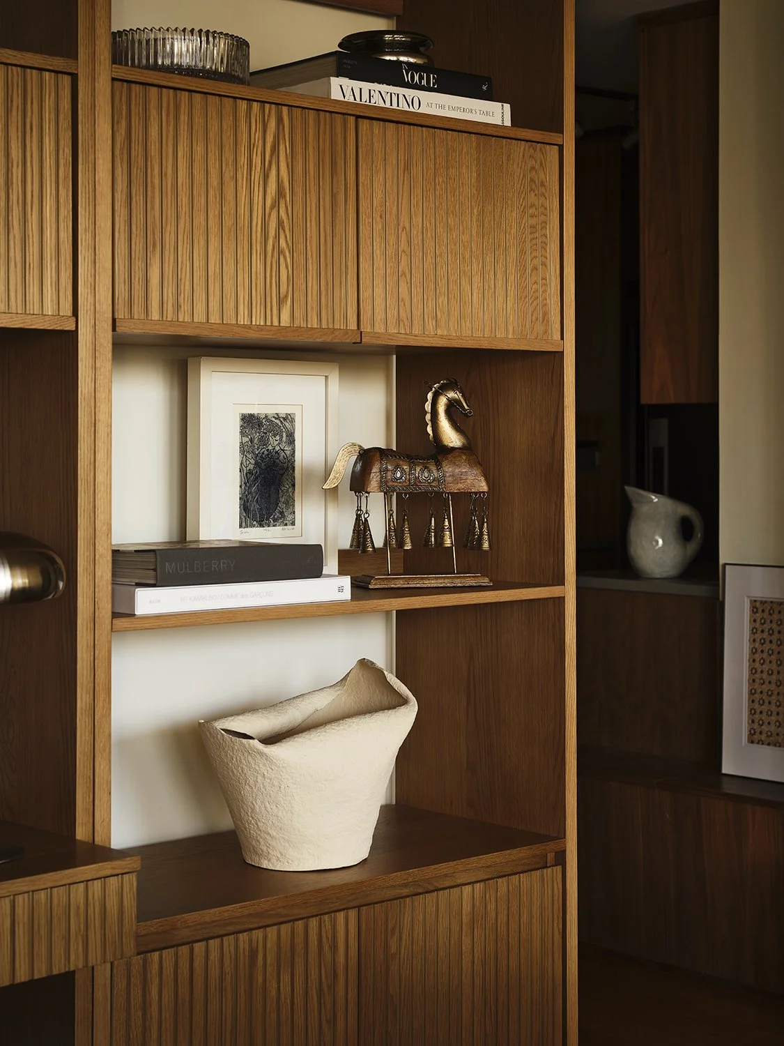 A wooden shelving unit with various decorative items including a ceramic bowl, a framed picture, a book titled 'Mulberry,' a brass horse figurine, and a vase.