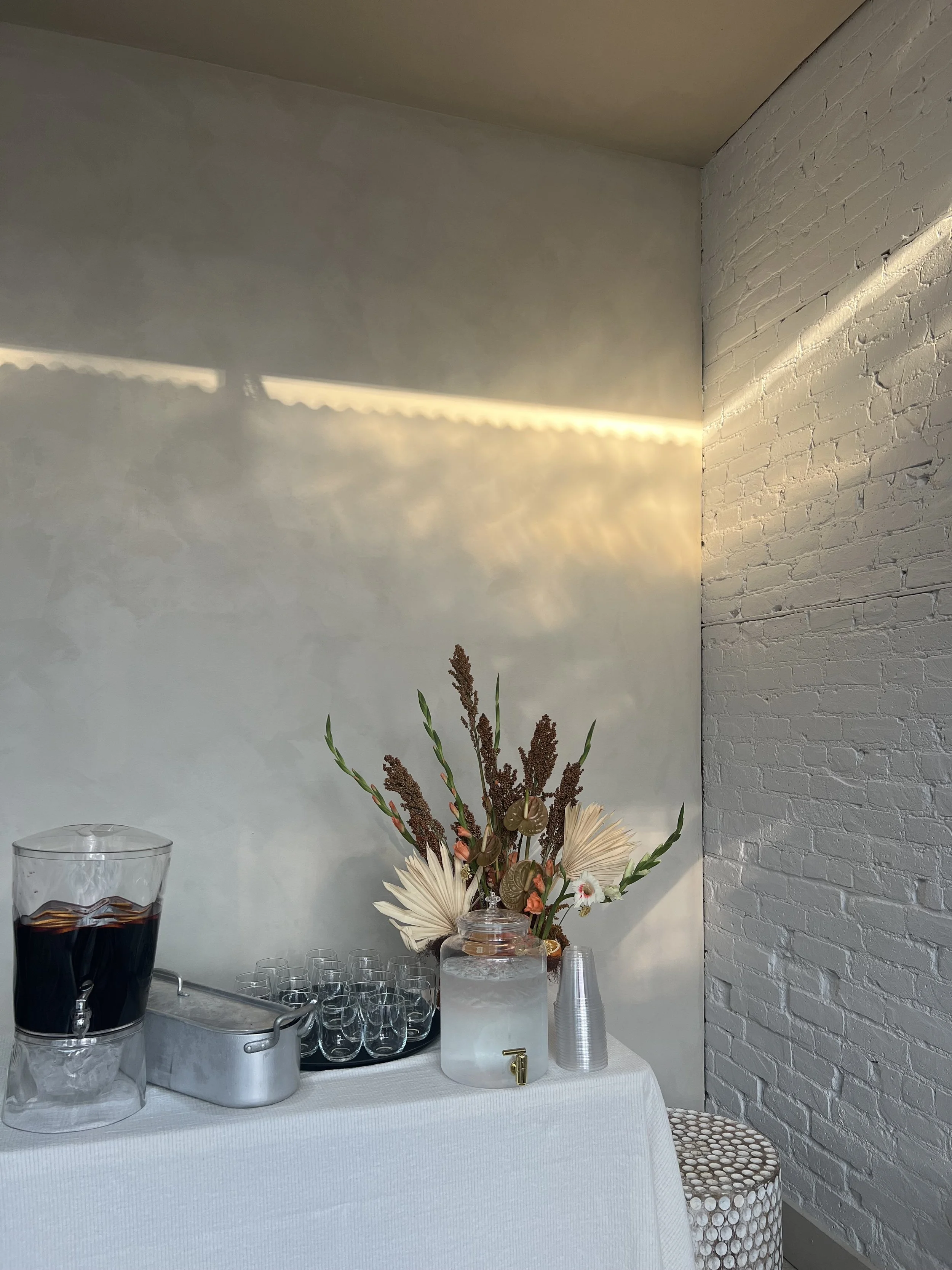 Decorative event space drink station with water dispenser, glasses, large floral arrangement, and disposable cups on a white table against a white brick wall and plain wall, illuminated by natural light.