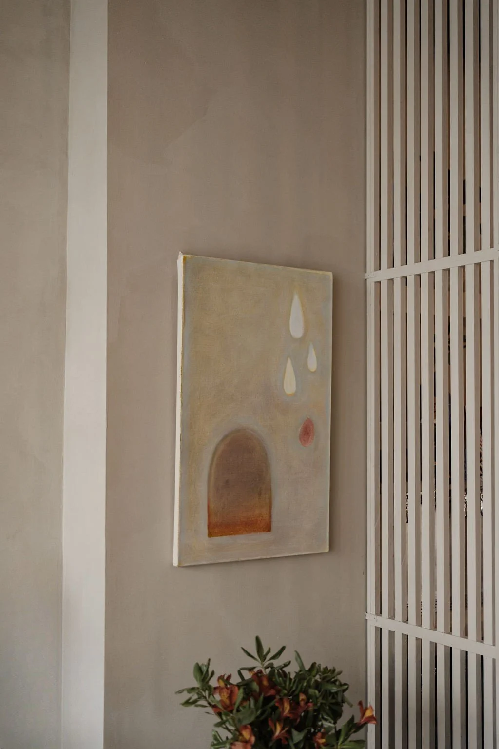A painting with abstract shapes hanging on a beige wall next to a slatted white wooden room divider and a plant with reddish leaves.