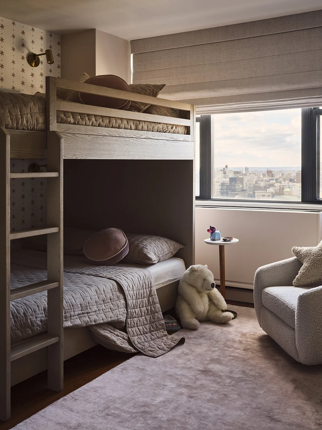 A bedroom with a loft bed, a plush teddy bear, and a cozy armchair near large windows overlooking a city skyline.