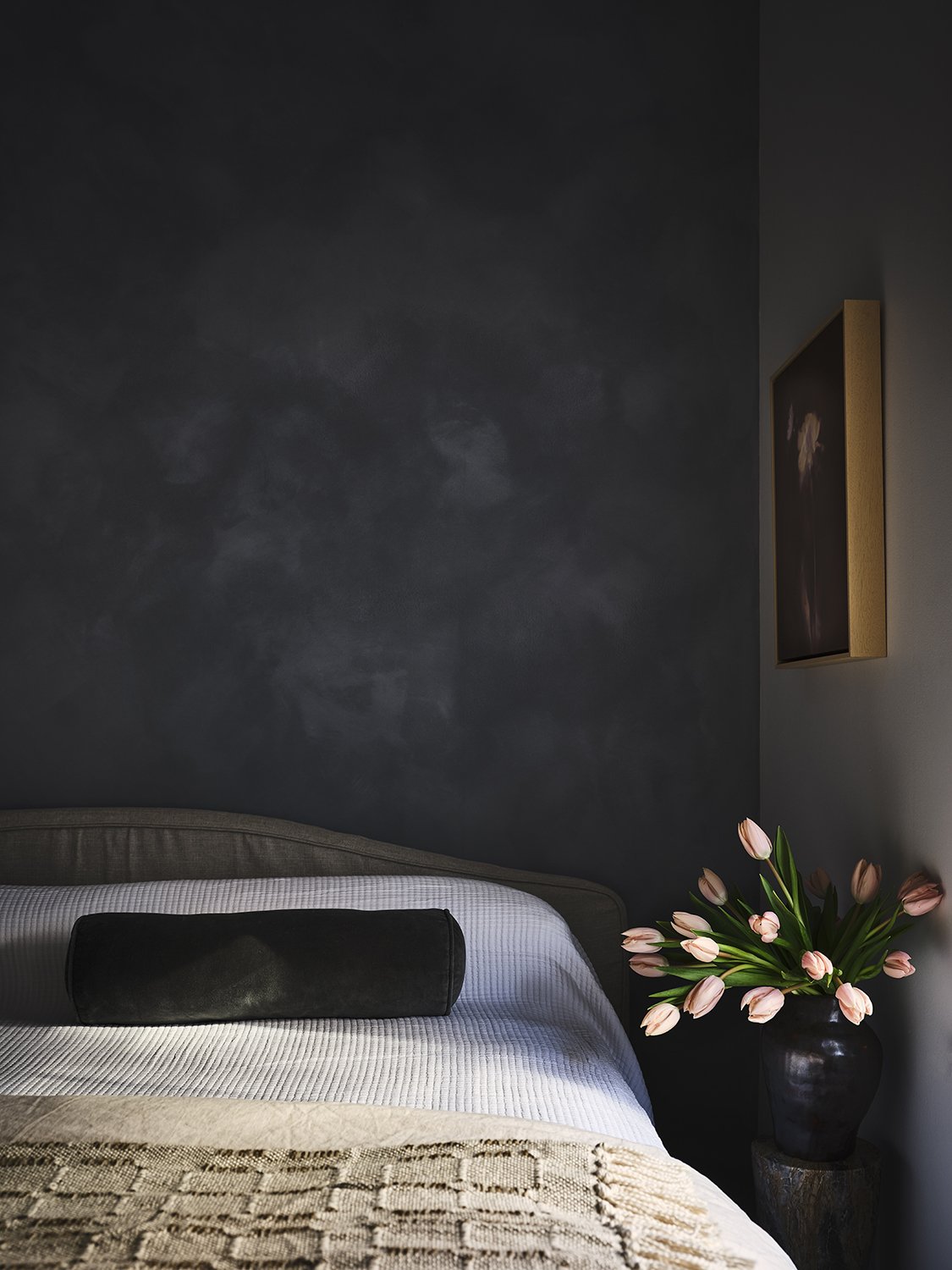 A bedroom with a bed featuring a textured beige blanket and gray pillows, a dark wall with a subtle pattern, a black vase with pink tulips on a small pedestal, and a framed painting on the wall.
