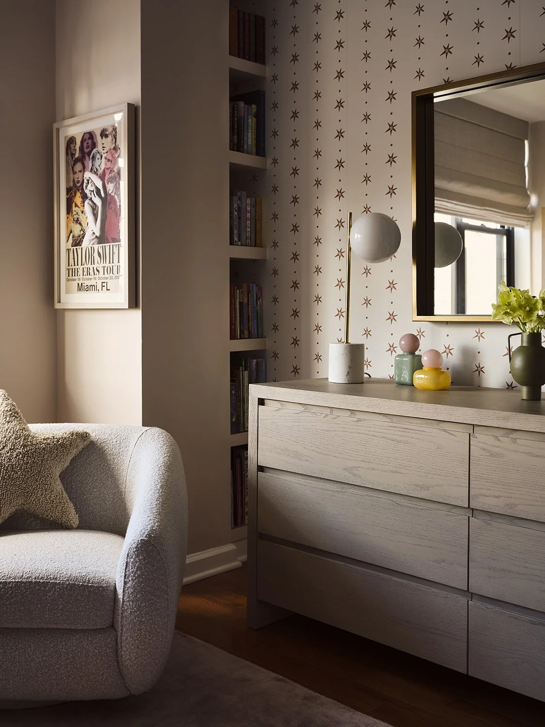 A cozy living room corner with a cream-colored armchair, a star-shaped pillow, a wooden dresser with decorative vases and a mirror, and a wall with star-patterned wallpaper and a built-in bookcase filled with books.