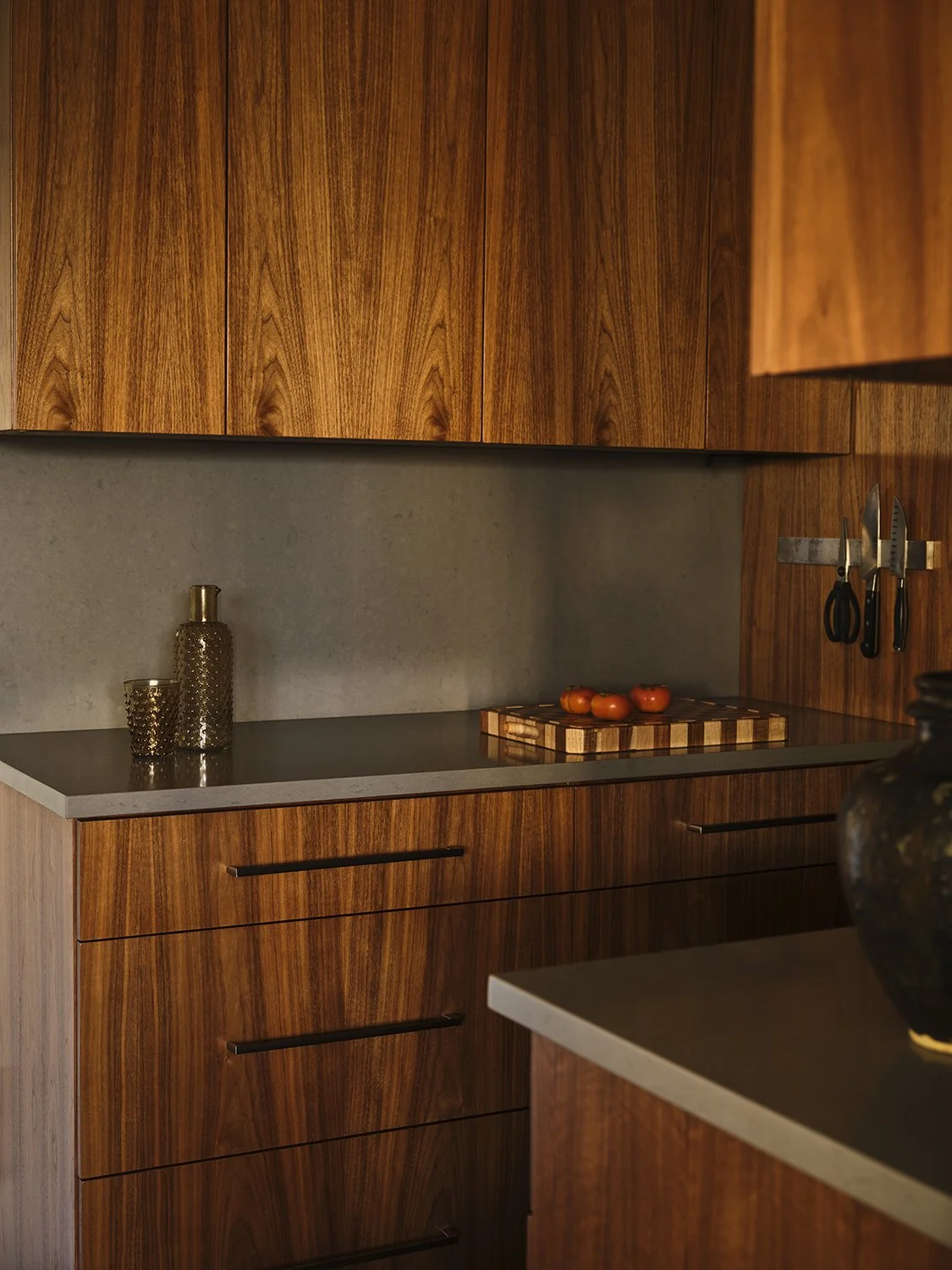 Modern kitchen with wooden cabinets, gray countertops, utensils on wall, tomatoes on cutting board, decorative jars on counter.