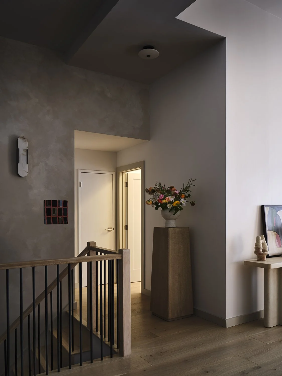 Interior view of a modern home with a staircase, a tall wooden pedestal with a floral arrangement, and minimalist art on the walls.