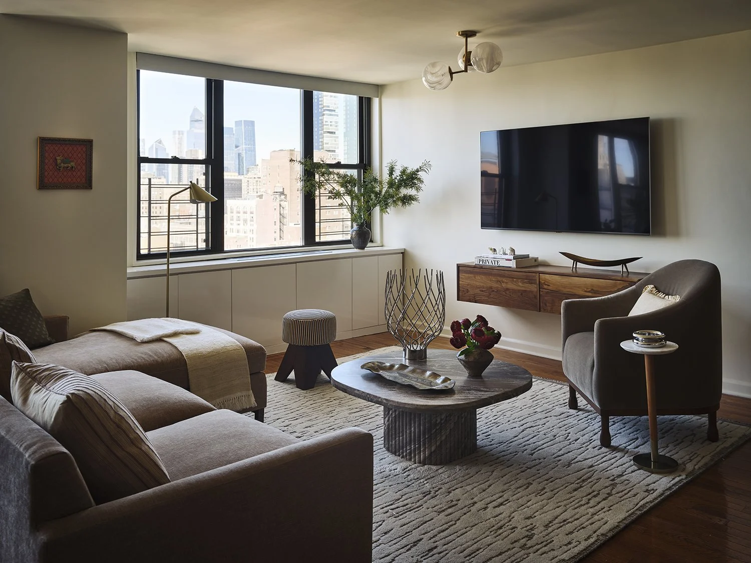 Living room with large window view of city skyline, beige sofa, armchair, wooden coffee table, wall-mounted TV, indoor plants, and modern decor.
