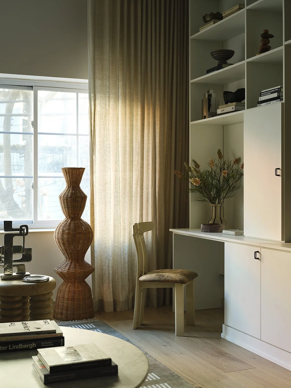 Living room corner with a window, beige curtains, a wooden chair, plants, books, and decorative objects.