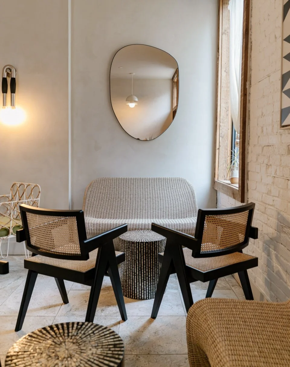 Cozy cafe seating area with two black armchairs, a beige textured sofa, and a small round patterned side table, set against a white brick wall with a window and a wall mirror.