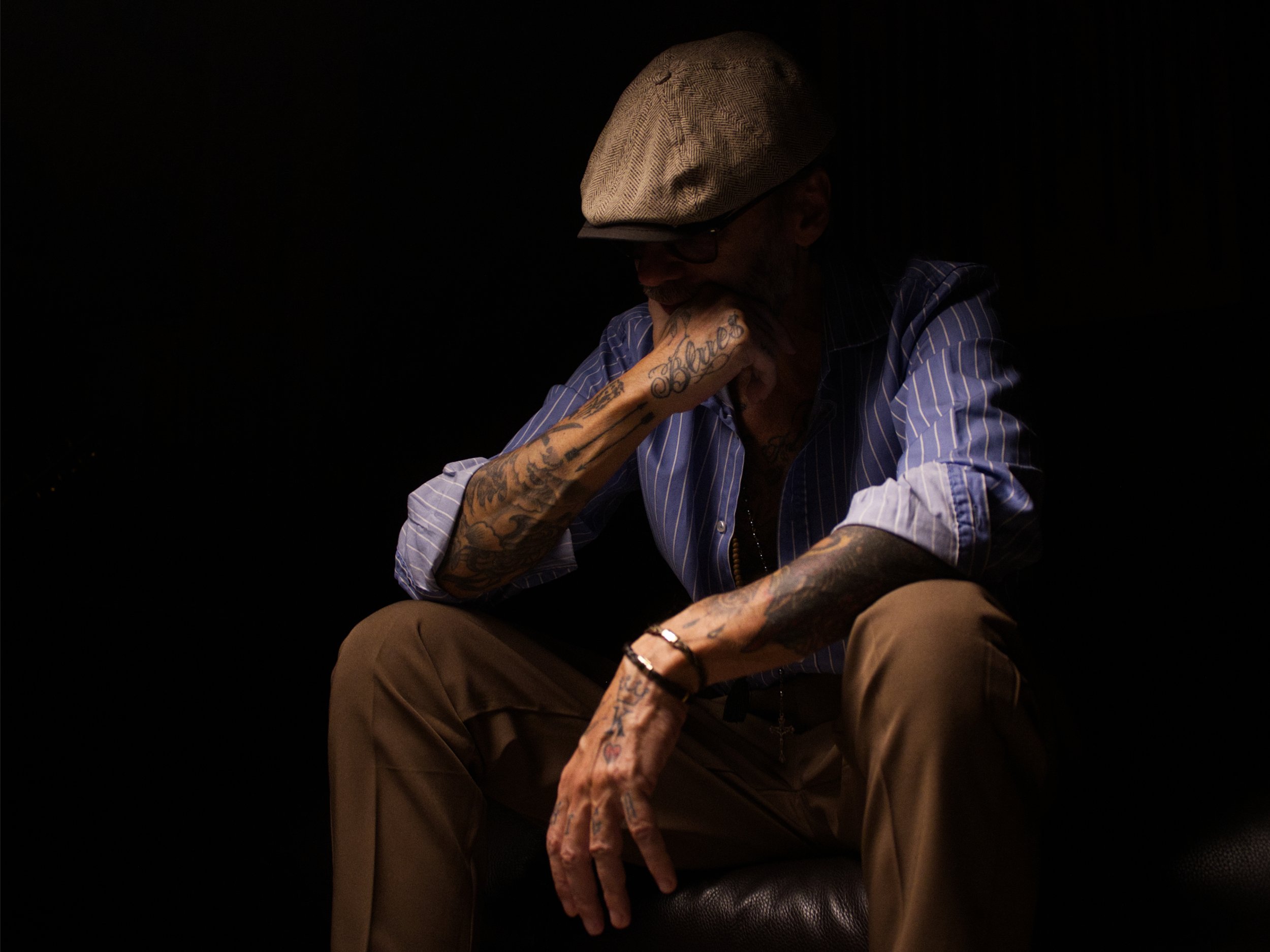 A man, bad dandy wearing a gray flat cap, glasses, a blue striped shirt with rolled-up sleeves, and khaki pants, sitting on a black surface in a dark setting, with his head resting on his hand.