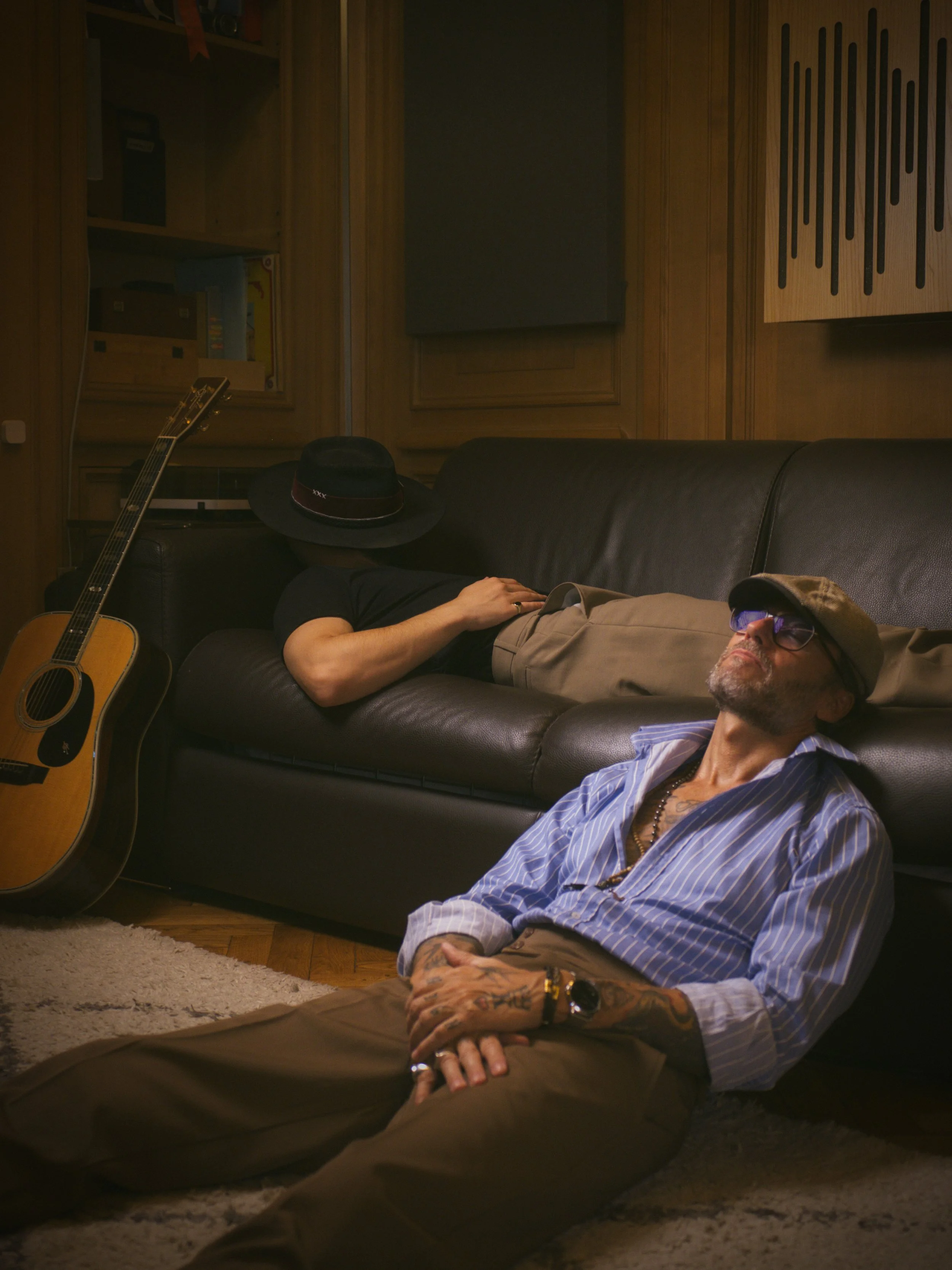 Two men resting in a music studio, one lying on the floor and the other on a black leather couch. The man on the floor has tattoos, glasses, a hat, a striped shirt, and beige pants. The man on the couch is partially covered by a hat and has dark hair and a black shirt. An acoustic guitar is leaning against the couch, and the room has wooden panels and soundproofing foam.