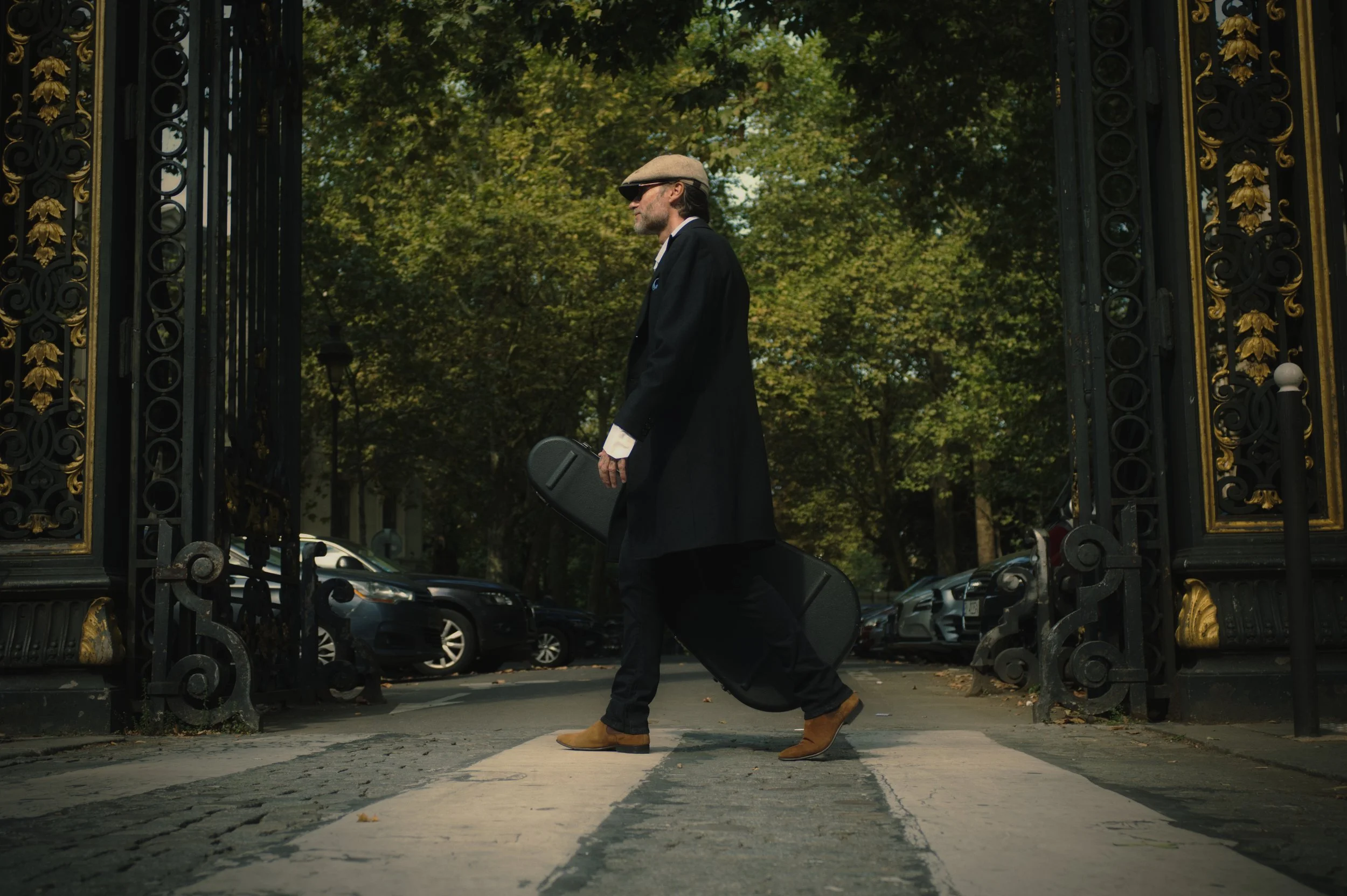 A man, BAD DANDY, walking across a pedestrian crosswalk carrying a guitar case, wearing sunglasses, a hat, a long dark coat, and brown shoes, with ornate black and gold gates open behind him and trees in the background.