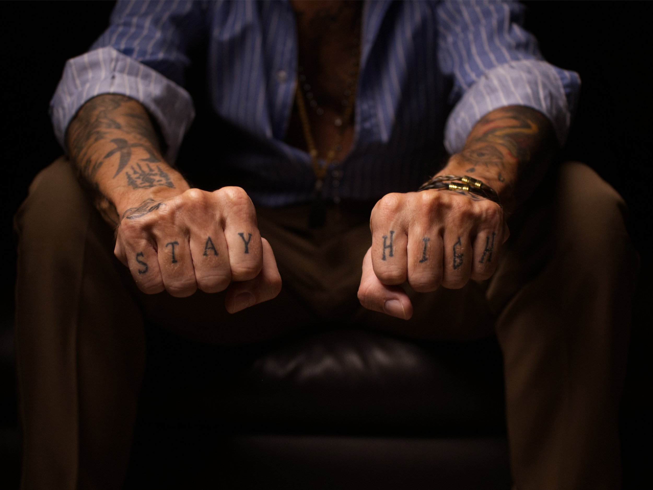Close-up of BAD DANDY with tattoos on his arms and hands, sitting with fists clenched. The knuckles spell out 'STAY HIGH' with tattooed letters.