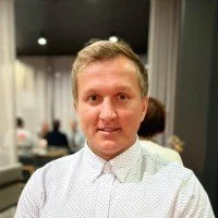 A man with short light-colored hair wearing a white button-up shirt, smiling in a well-lit indoor setting with blurred people in the background.