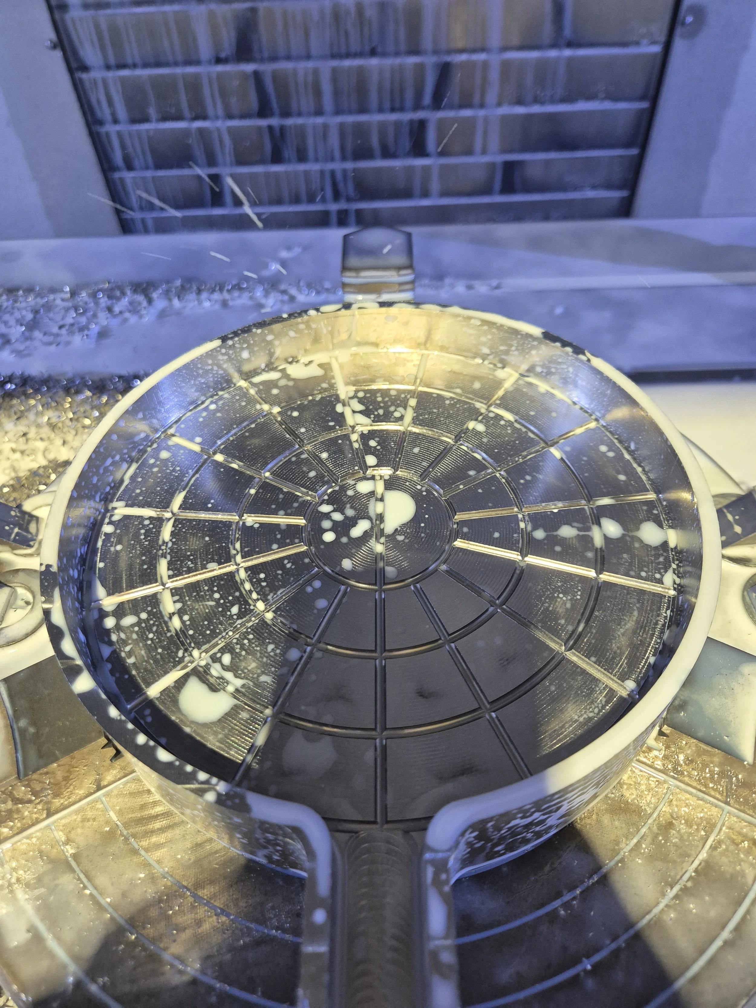A metal waffle maker with batter inside, sitting on a stovetop stovetop with a kitchen window in the background.