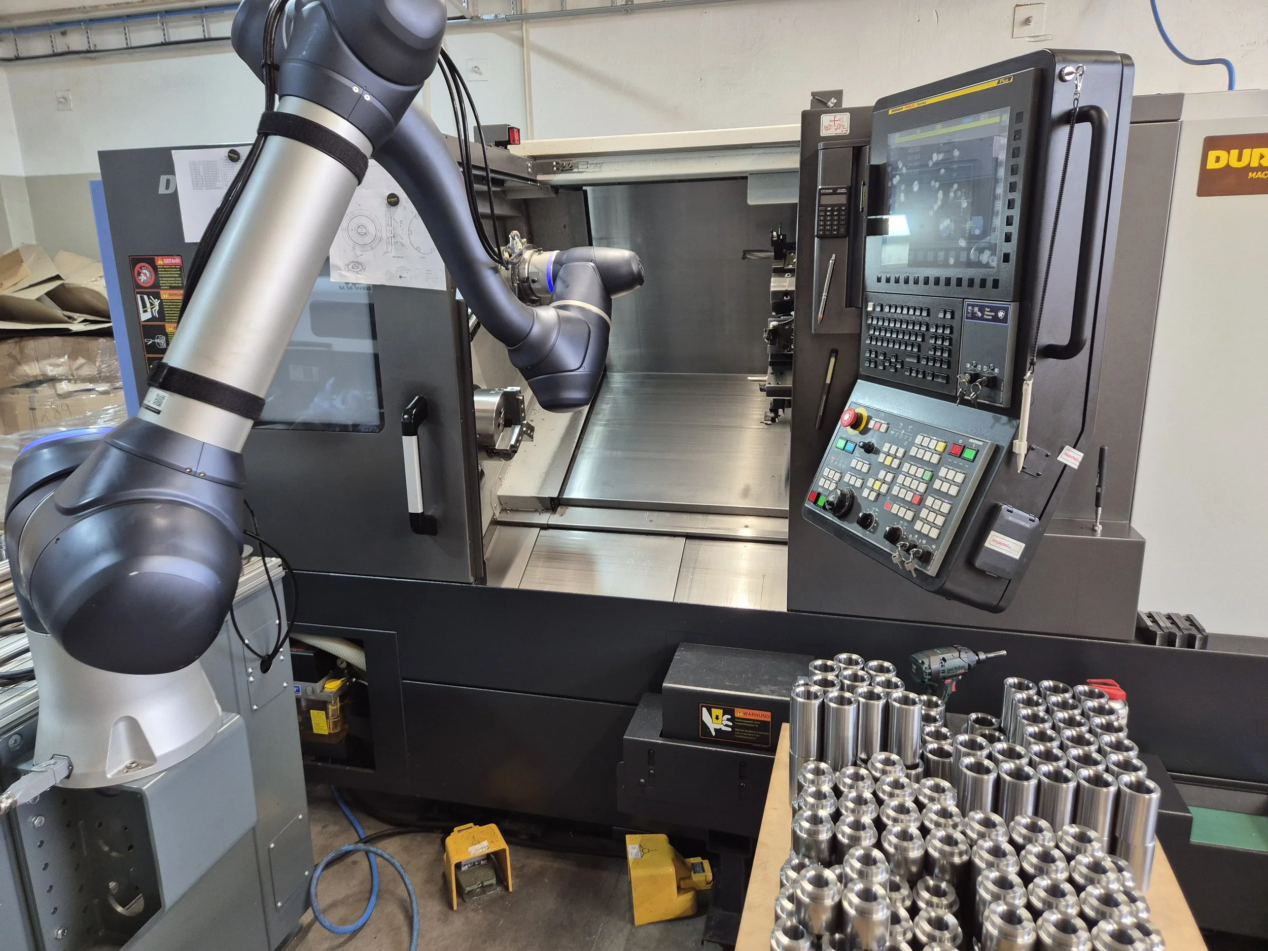 A CNC machining center with a robotic arm, control panel, and multiple metal cylindrical parts on a table.