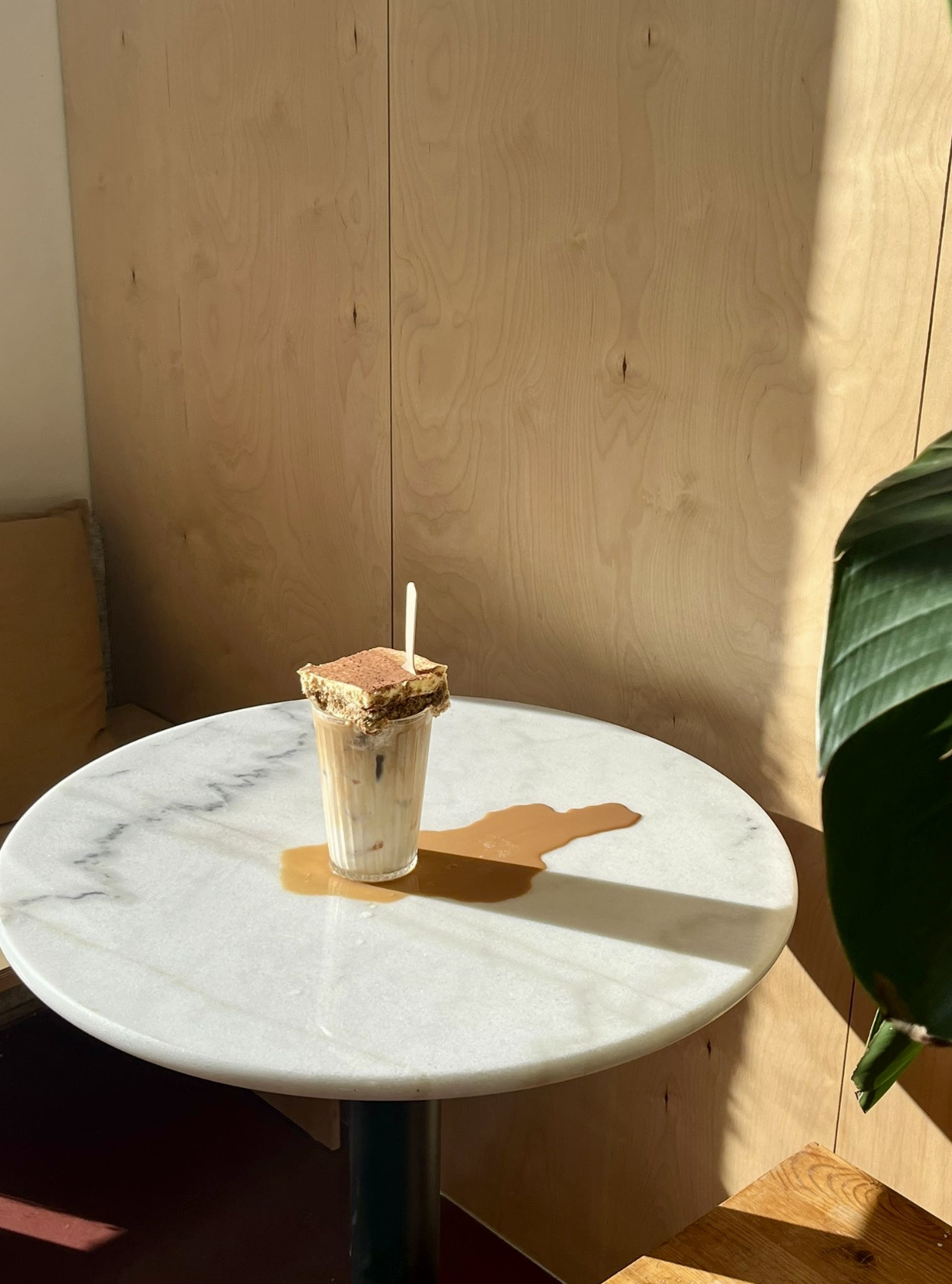 Tiramisu latte, sobre uma mesa de mármore branca, com sombra visível e uma parede de madeira ao fundo e uma planta à direita.
