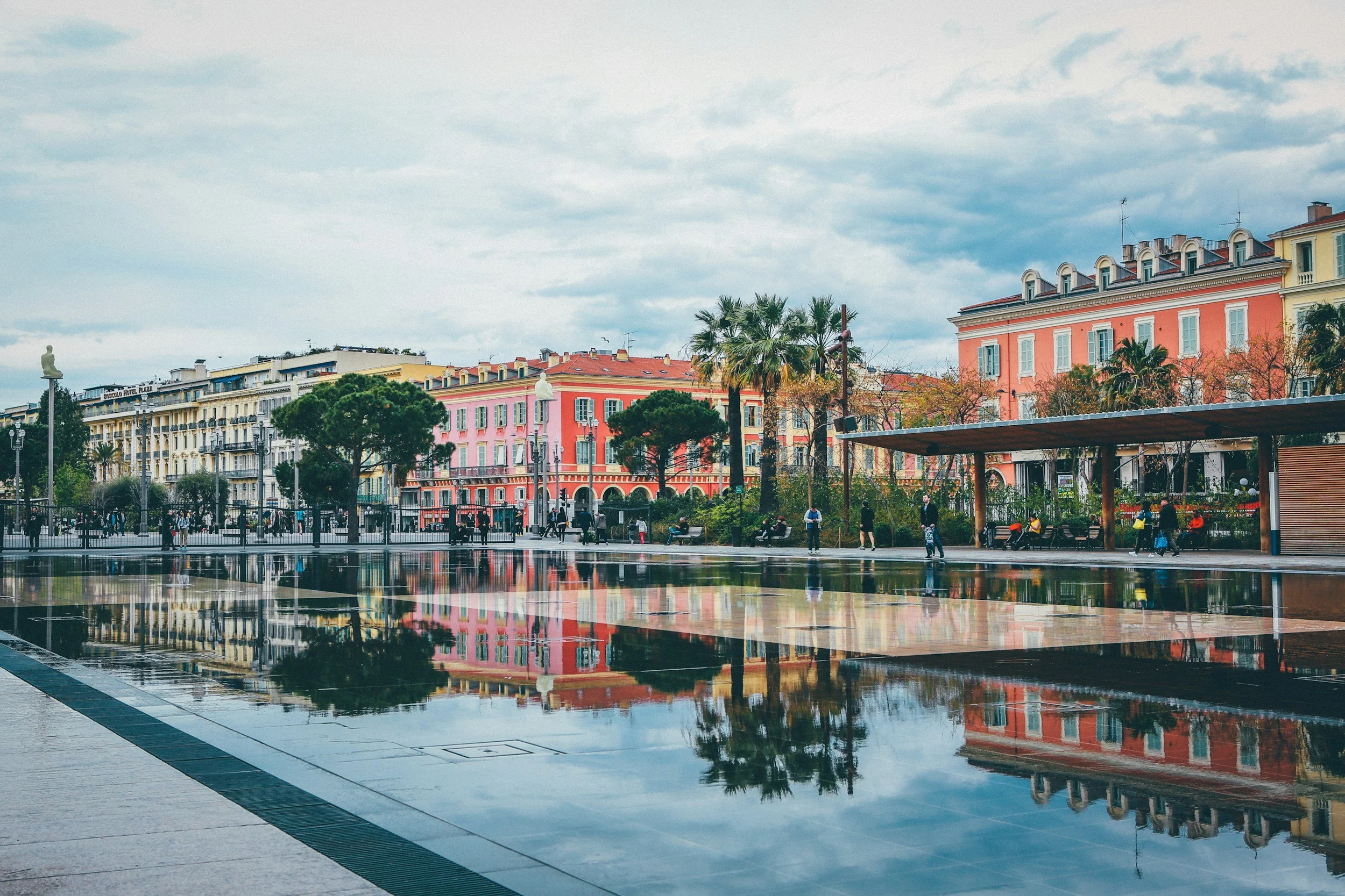 City square with colorful buildings, trees, benches, and a reflective water feature, with people walking and sitting, under a cloudy sky.