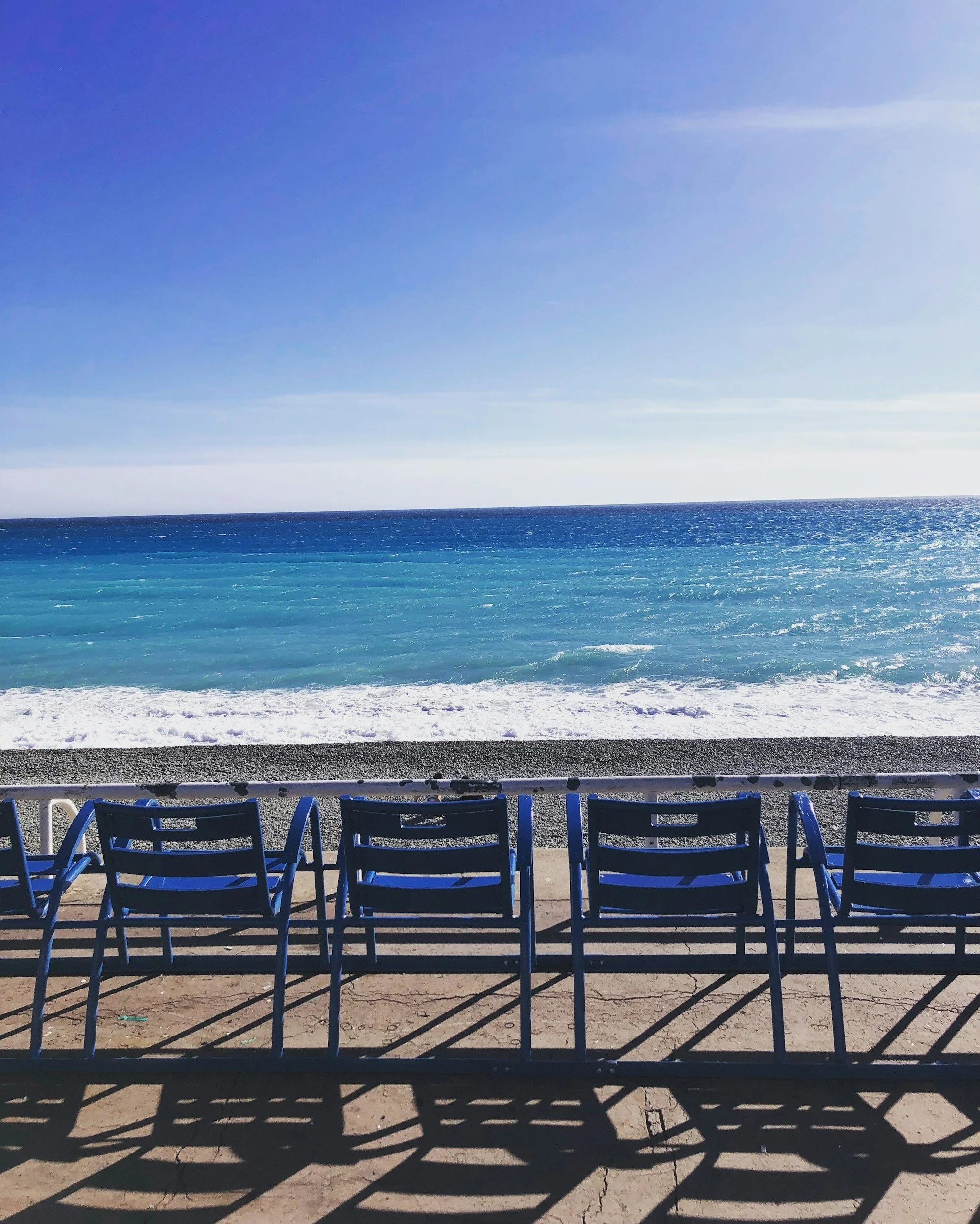 Blue chairs lined up on a concrete sidewalk facing the ocean, with waves crashing and a clear sky overhead