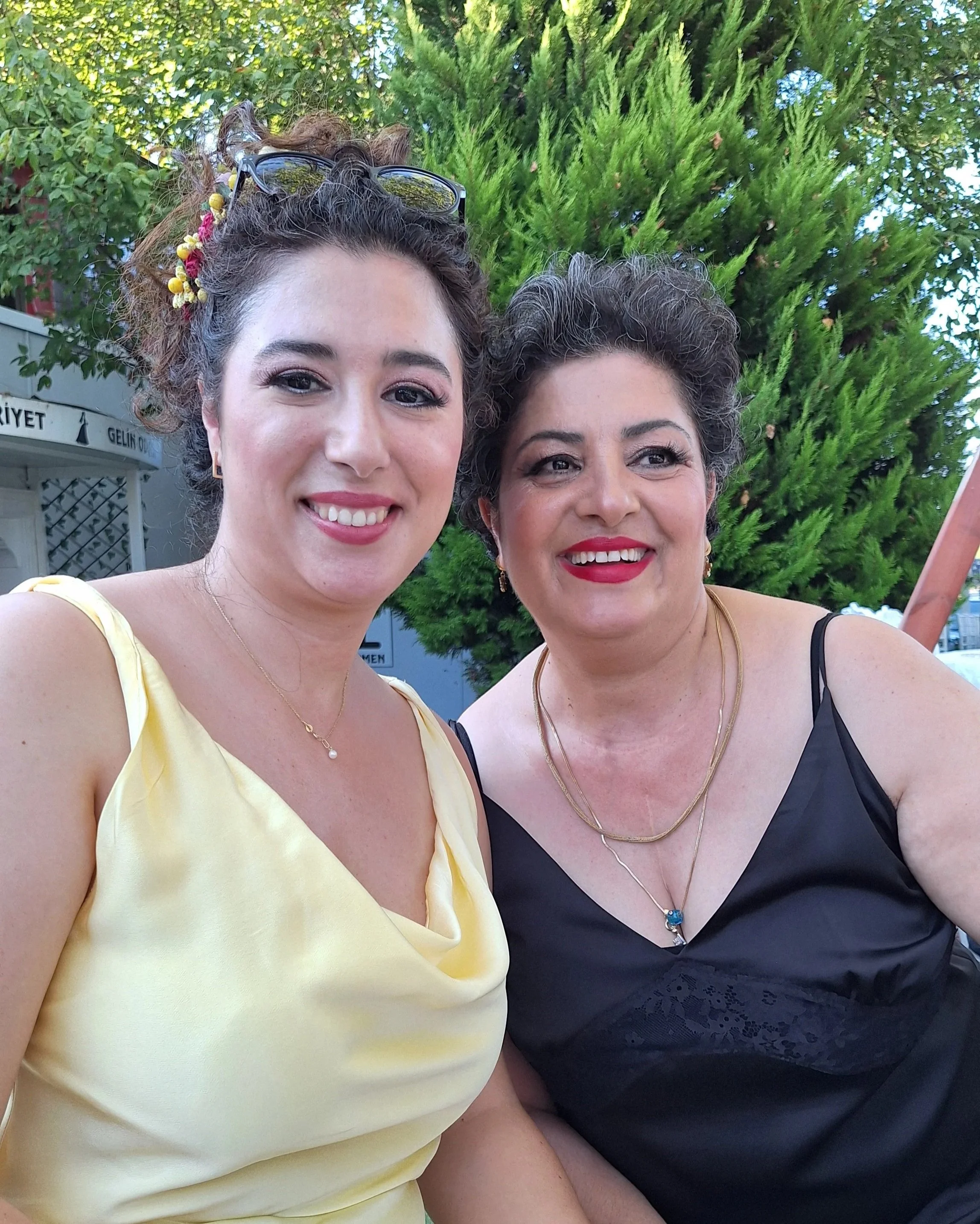 Two women smiling outdoors with green trees in the background, one wearing a yellow dress and the other a black dress, both with jewelry and accessories.