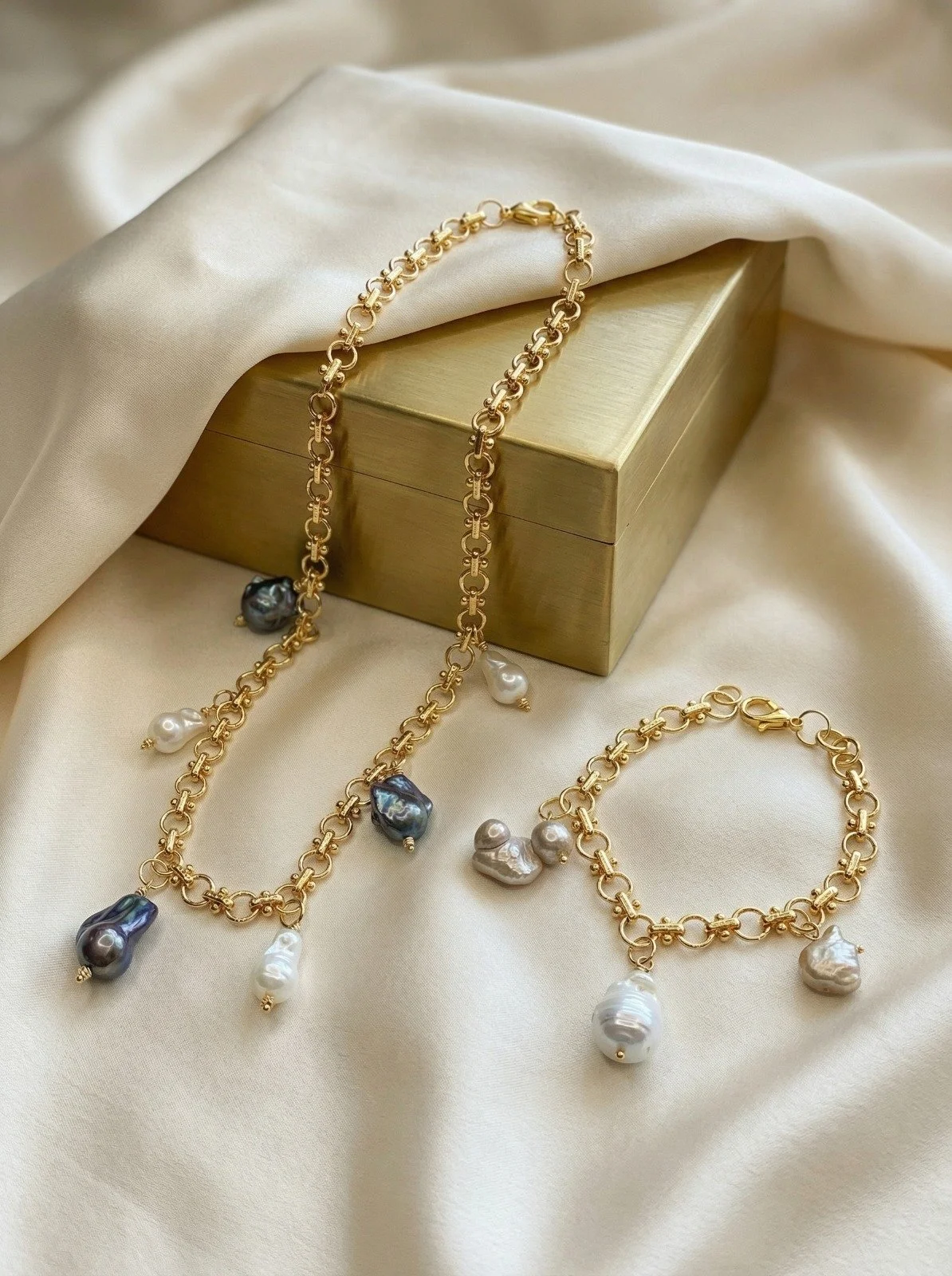 Gold jewelry set of a necklace and bracelet with dangling pearls and baroque pearls on silk fabric beside a gold-colored box.