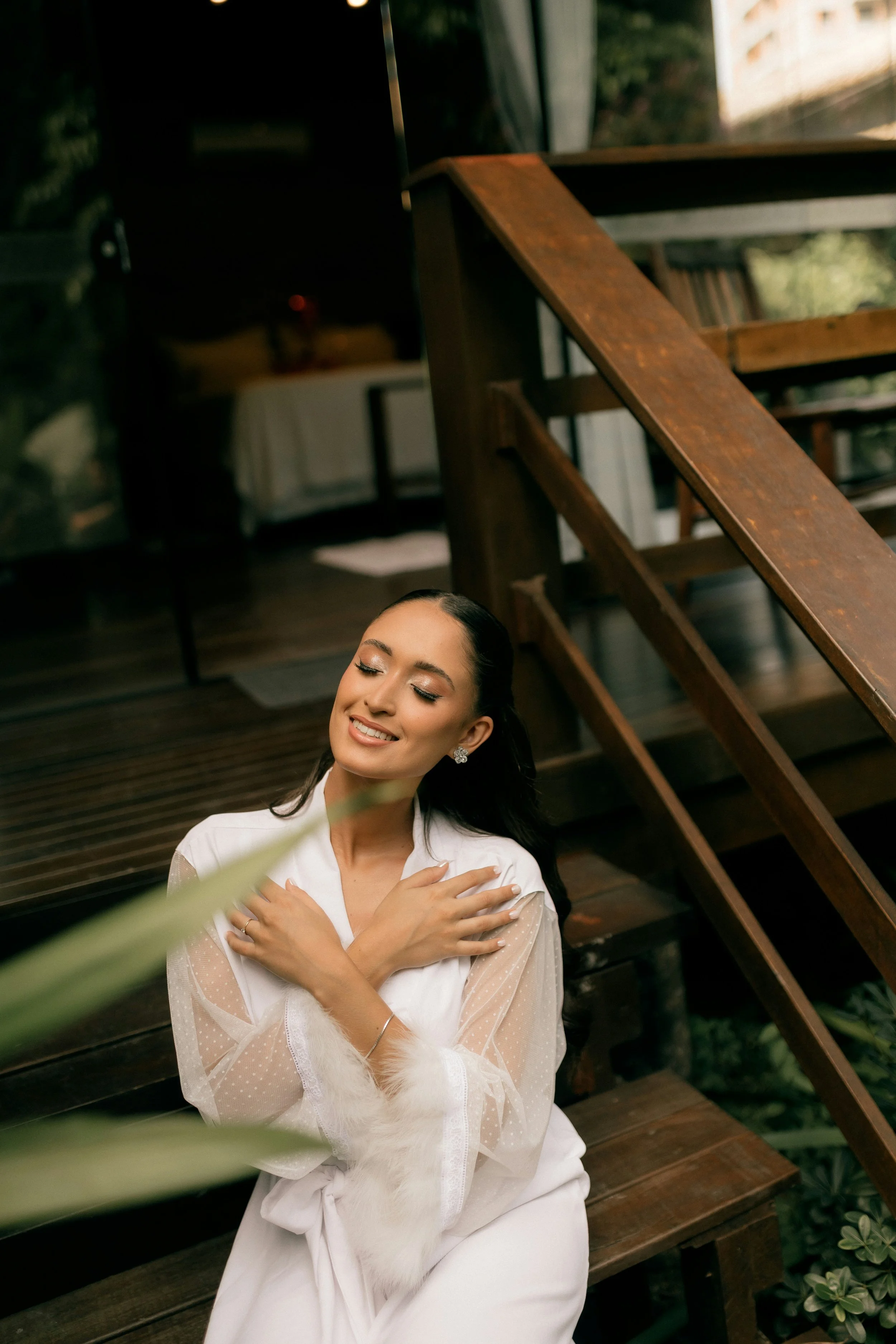 A woman with dark hair and earrings, wearing a white dress with sheer sleeves, sitting outdoors on wooden stairs near greenery, with a serene expression and her arms crossed over her chest.