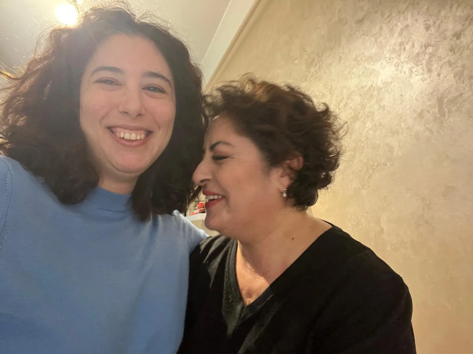 Two women smiling and leaning close to each other, standing indoors near a beige wall with a textured pattern.