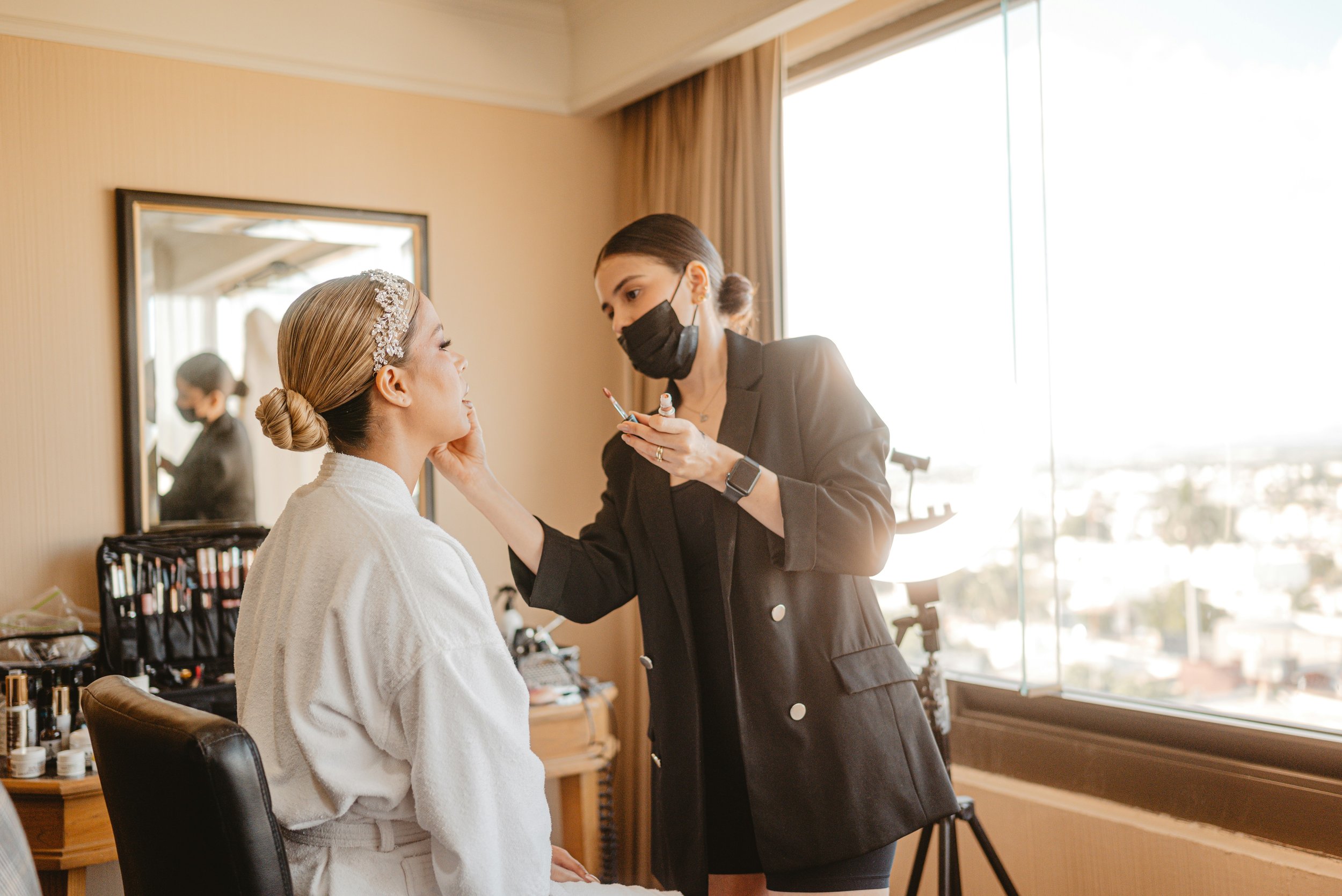 A woman seated in a makeup chair receives makeup application from a makeup artist wearing a black face mask in a well-lit room with a large window and a mirror.