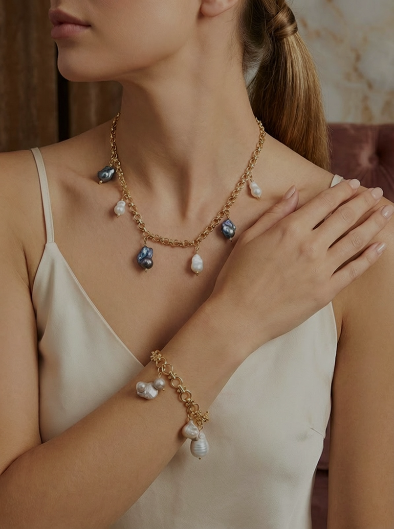 A woman wearing a gold chain necklace with black and white pearl pendants and a matching pearl bracelet, with her left hand resting on her right shoulder.