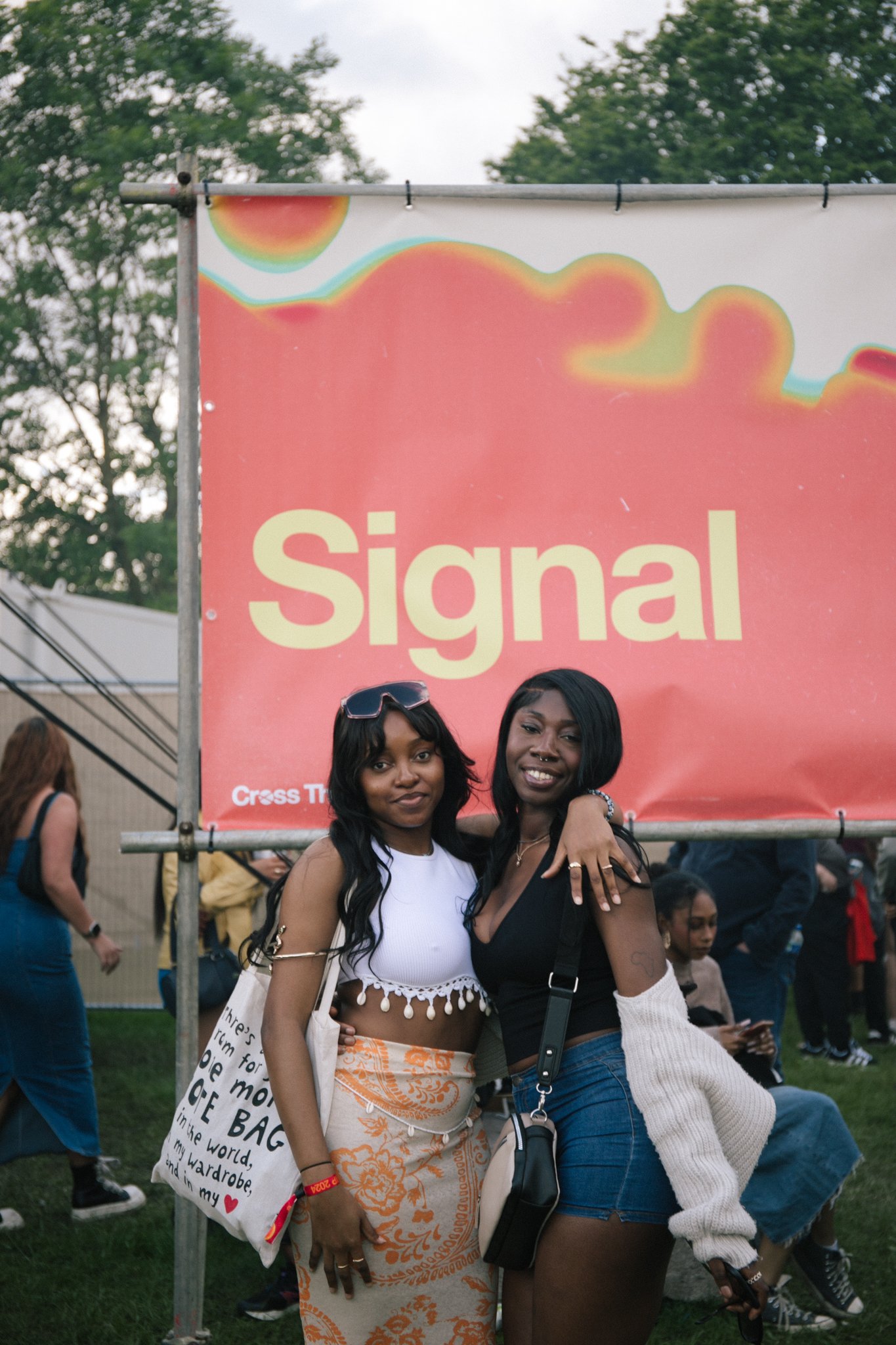 Two women standing close together with their arms around each other, smiling outdoors at a gathering or festival, with a pink sign in the background that says 'Signal' and has colorful design elements.