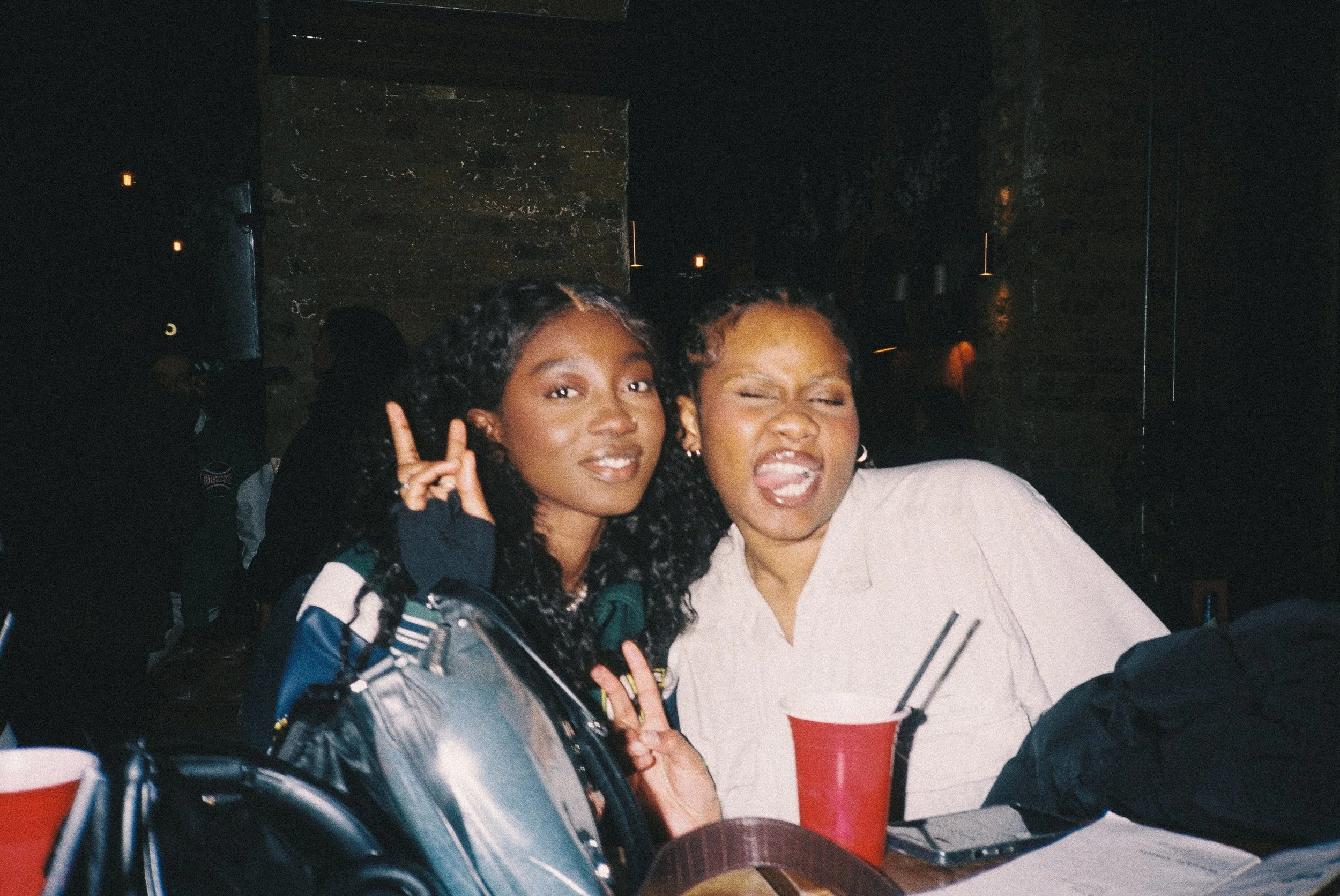 Two young women making playful faces at a social gathering. One girl is holding up a peace sign expression, and the other is sticking out her tongue. They are sitting at a table with red cups. The background is dark with some dim lighting.
