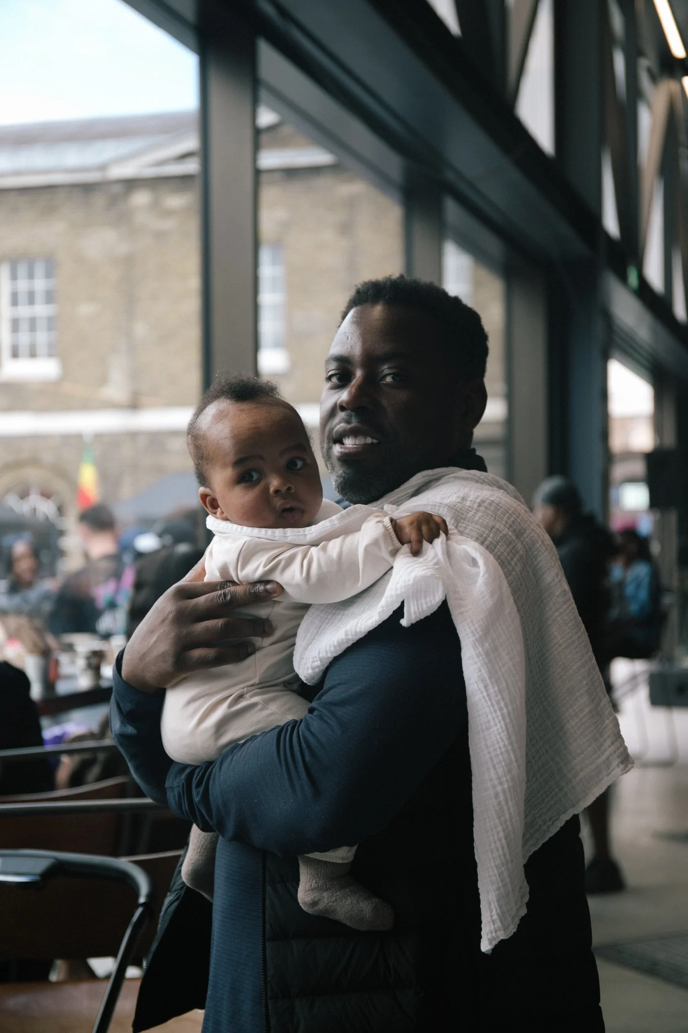 A man with dark skin holding a baby with dark skin in an indoor public space with large windows and blurred background of people and buildings.
