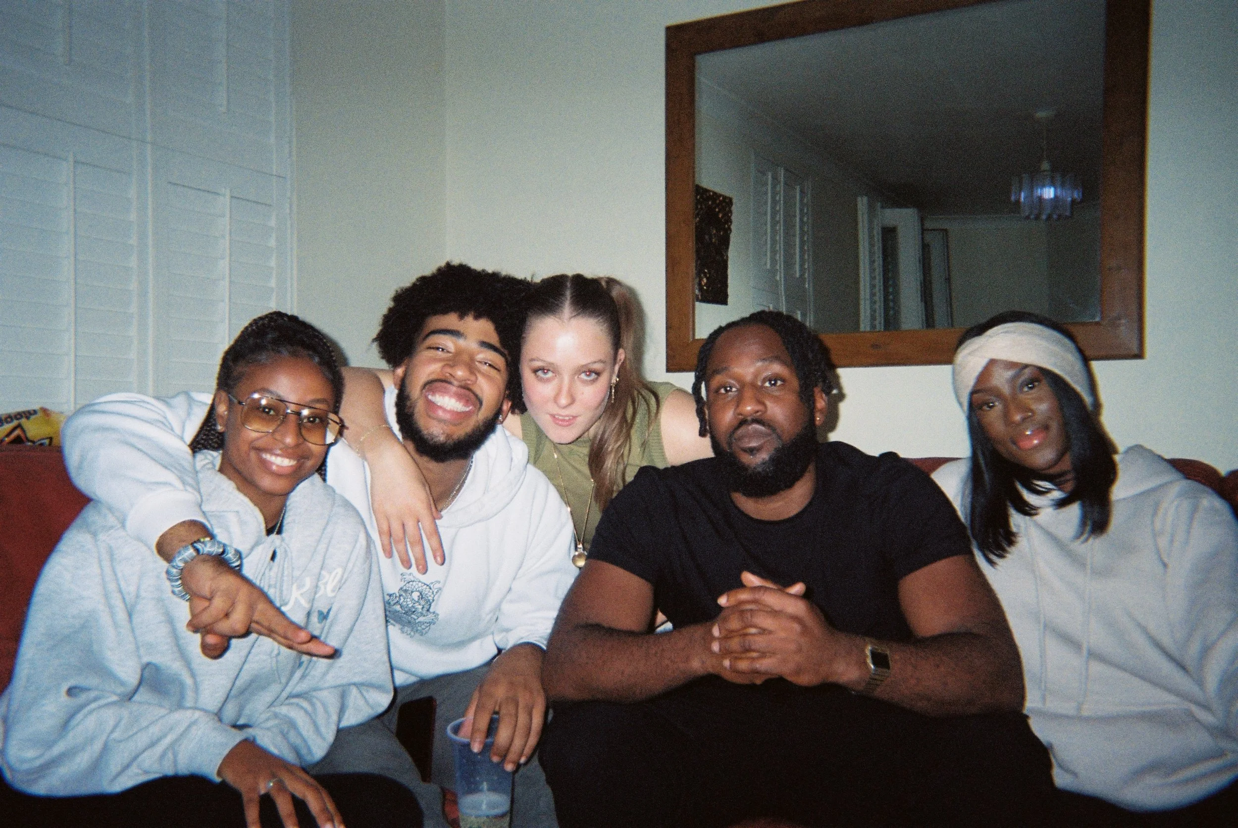 A group of five diverse friends sitting closely together on a couch, smiling and enjoying each other's company in a cozy living room with a mirror and a chandelier in the background.