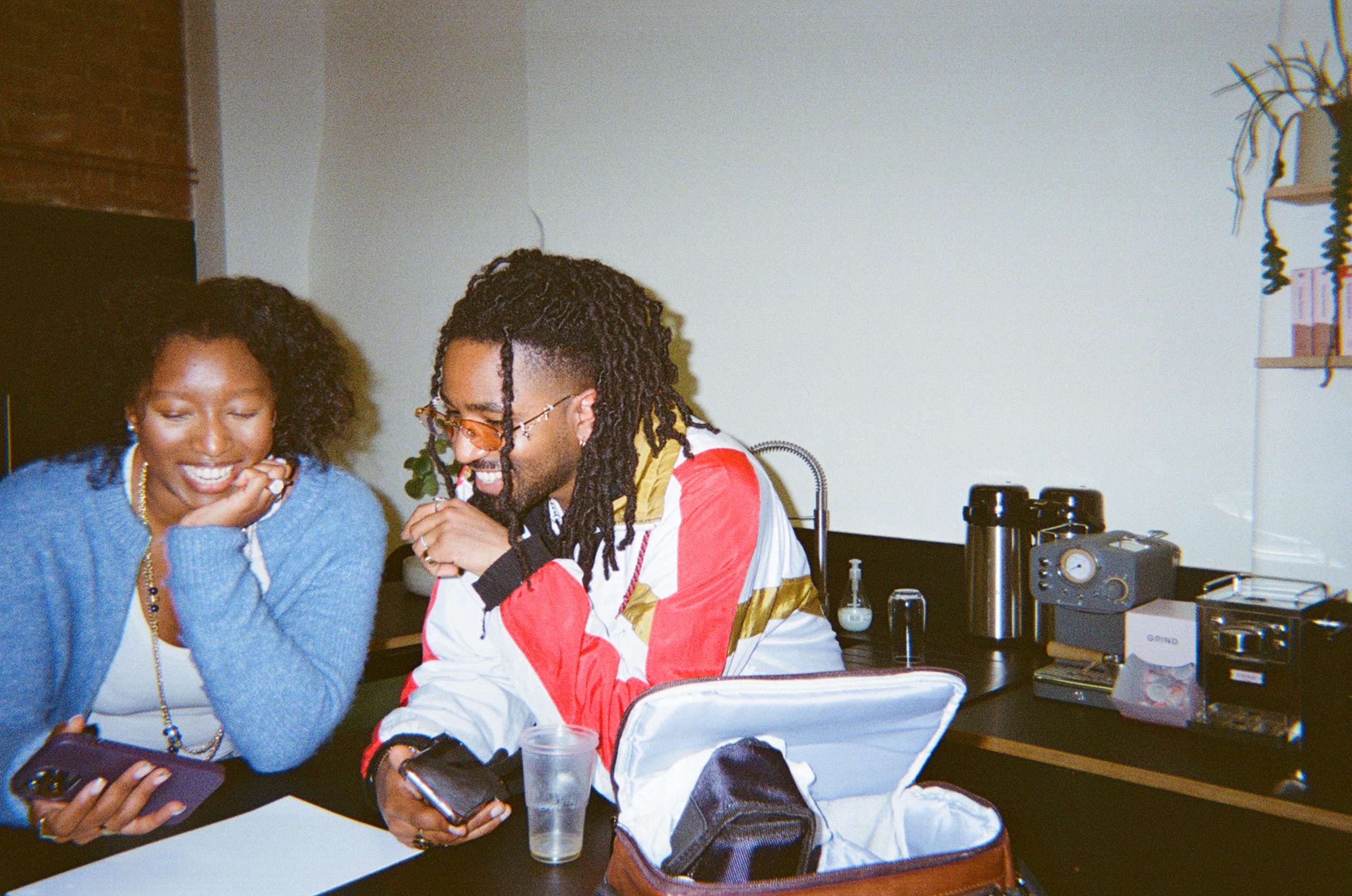 A man with dreadlocks and sunglasses and a woman with curly hair smiling and looking at their phones in an indoor setting with scientific equipment and supplies on the table.