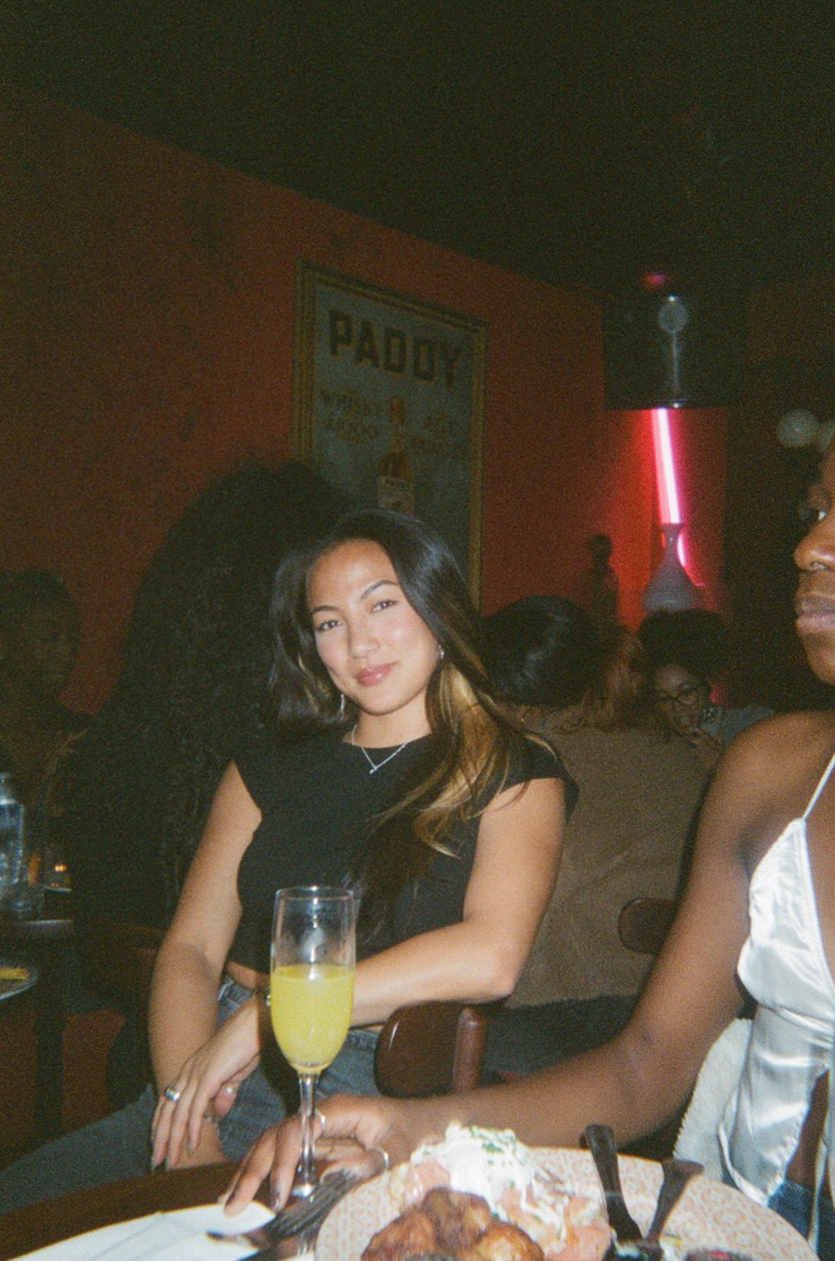 A young woman with long dark hair and a black top sitting at a table in a dimly lit restaurant or bar with red walls, a poster on the wall, and other patrons in the background. There is a glass of yellow drink and food on the table.