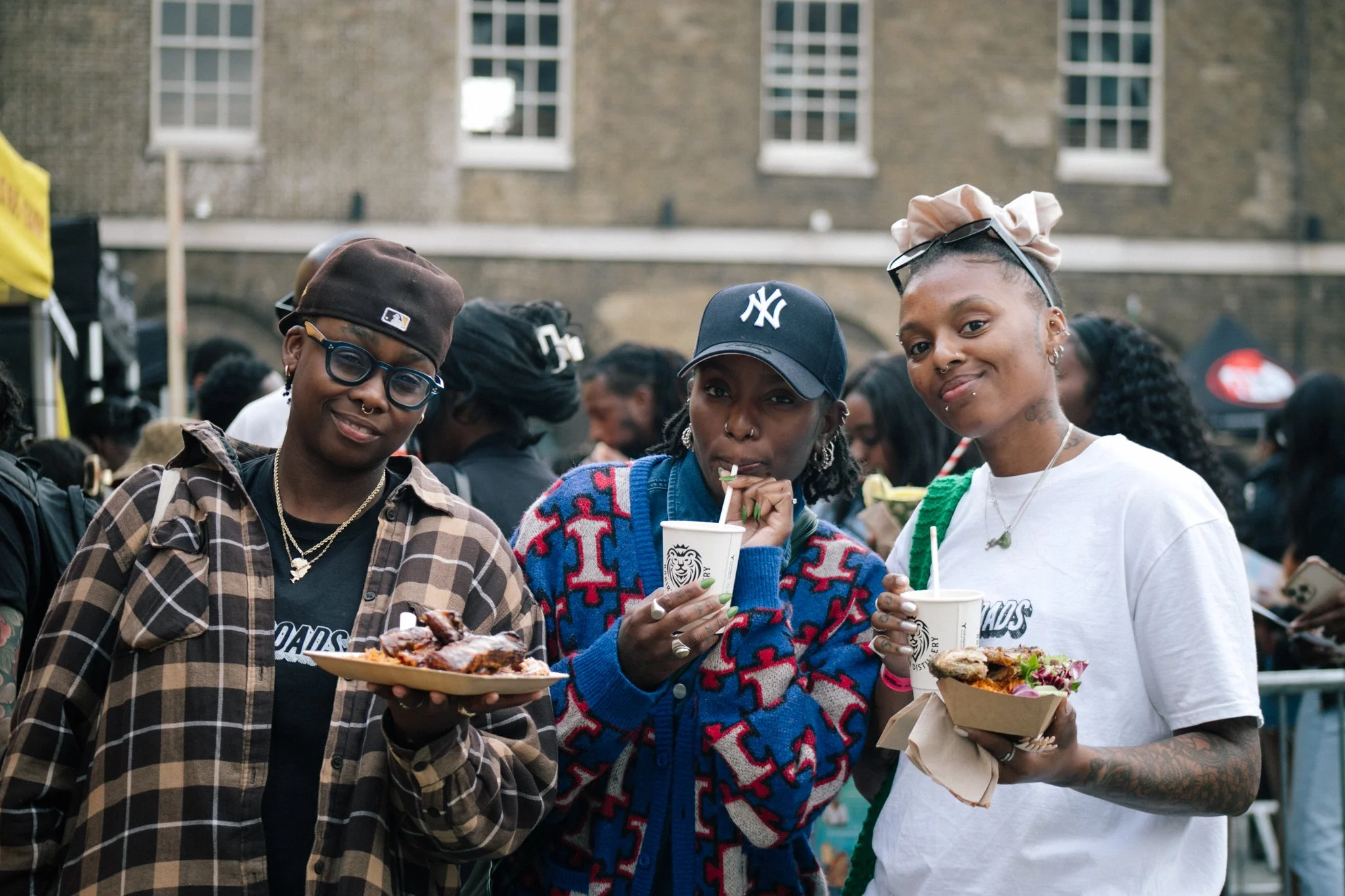 A crowd of people at an outdoor event. Three women in the foreground are holding plates and cups with food and drinks, smiling and enjoying each other's company.