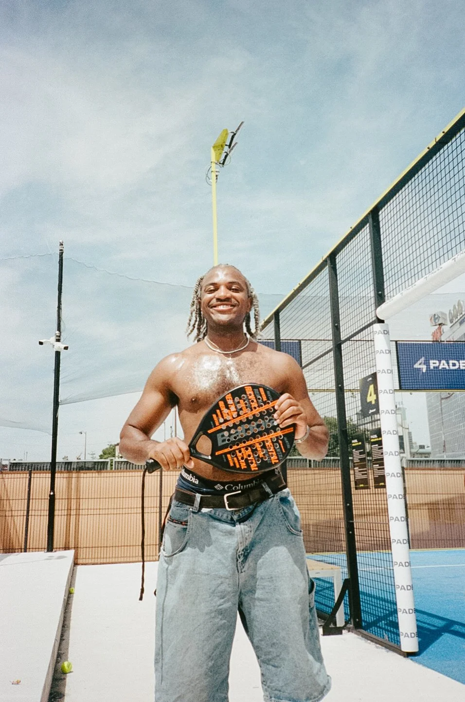 A man with grey hair in twists smiling and holding a padel racket on a padel court outdoors.