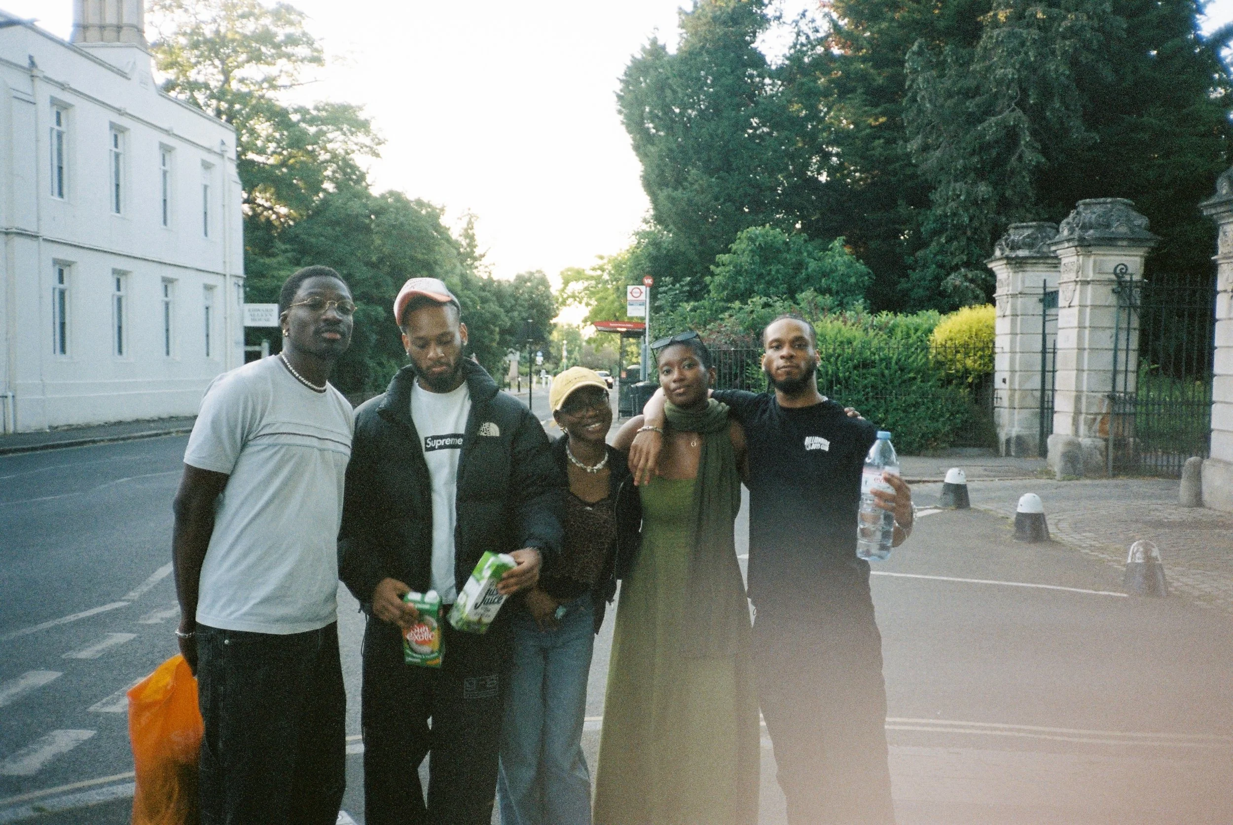 Group of five young adults standing together outdoors on a city street, with trees and historic building entrance in the background, during late afternoon or early evening.