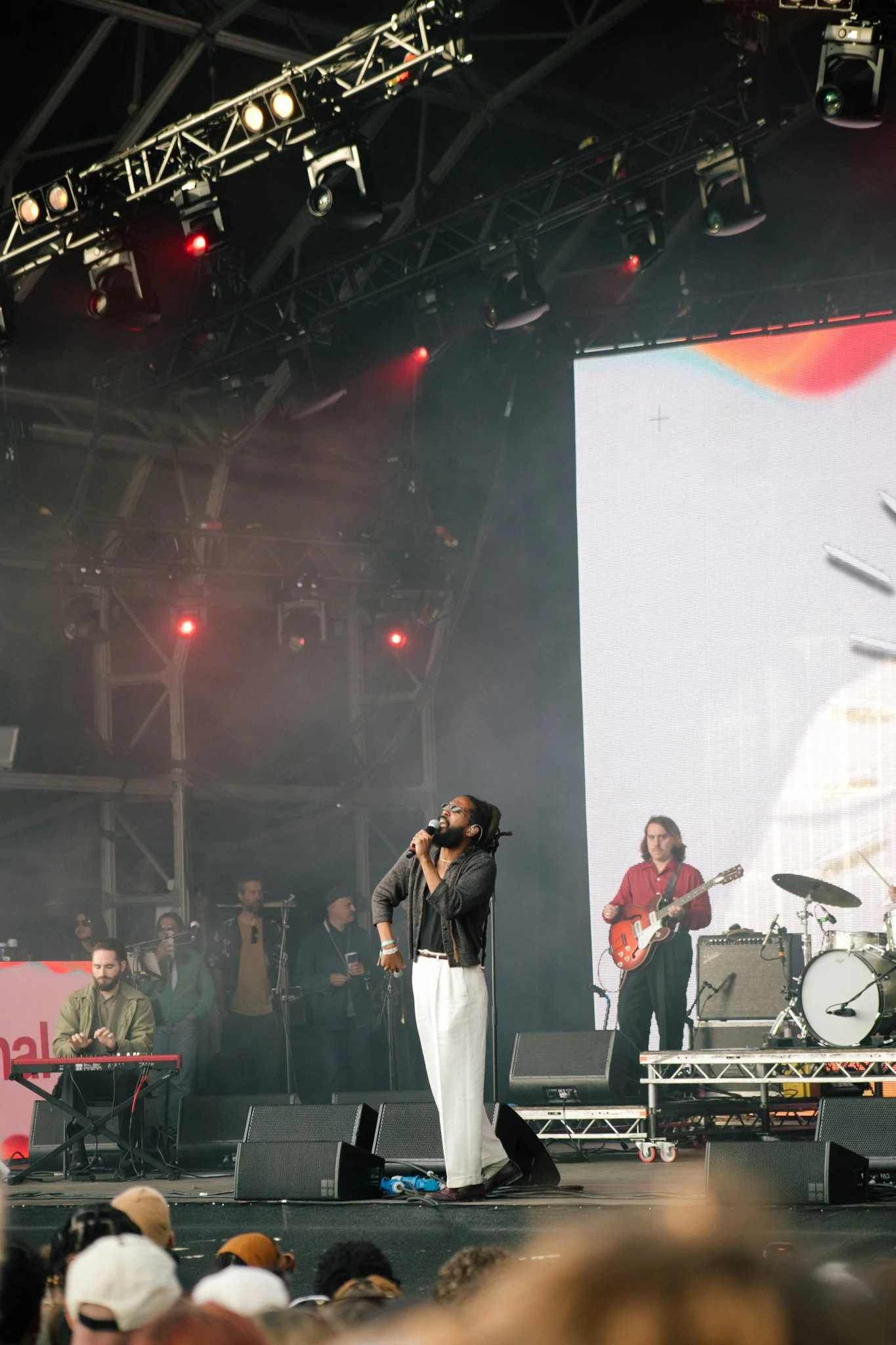 A live outdoor concert with musicians  Thee Sacred Souls on stage, including a man with long dreadlocks singing into a microphone and a guitarist playing an electric guitar. There is a crowd in front watching the performance.