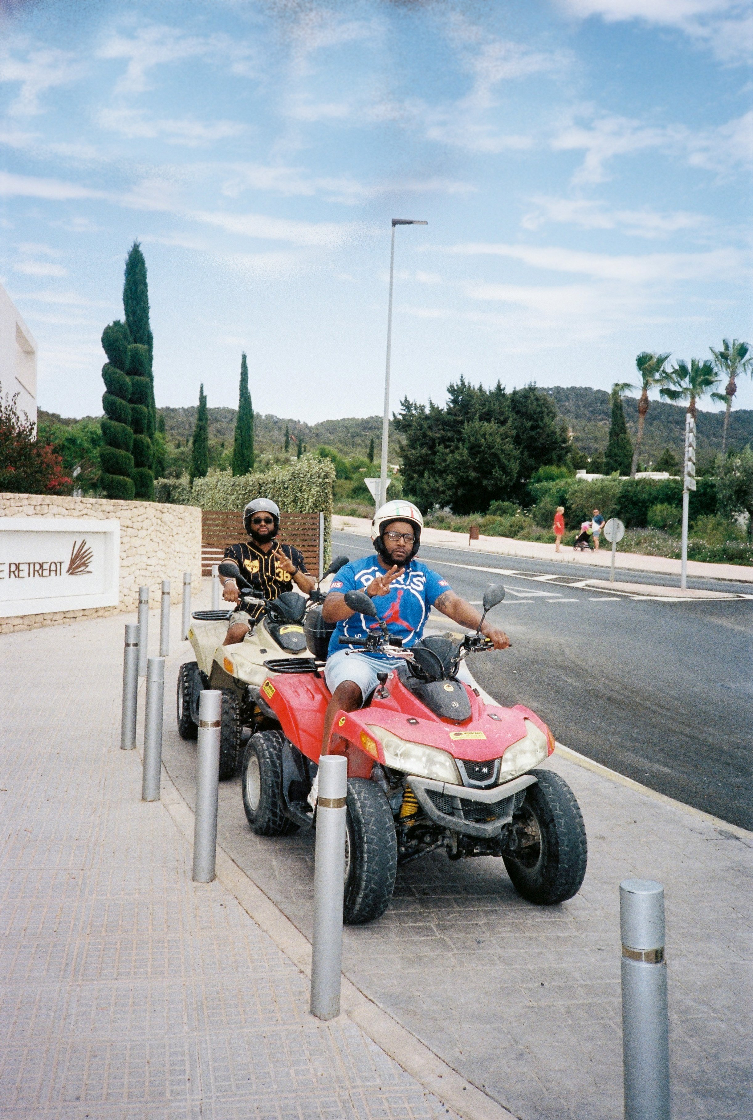 Two men riding all-terrain vehicles (ATVs) on a sidewalk in a sunny outdoor setting. One man is wearing a black and yellow shirt and the other a blue shirt with various logos. They are wearing helmets and sunglasses.