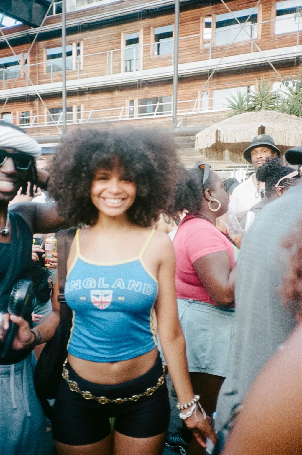 A young woman with curly hair smiling at a gathering, wearing a blue England tank top, black shorts with a chain belt, and jewelry, surrounded by other people at an outdoor event.
