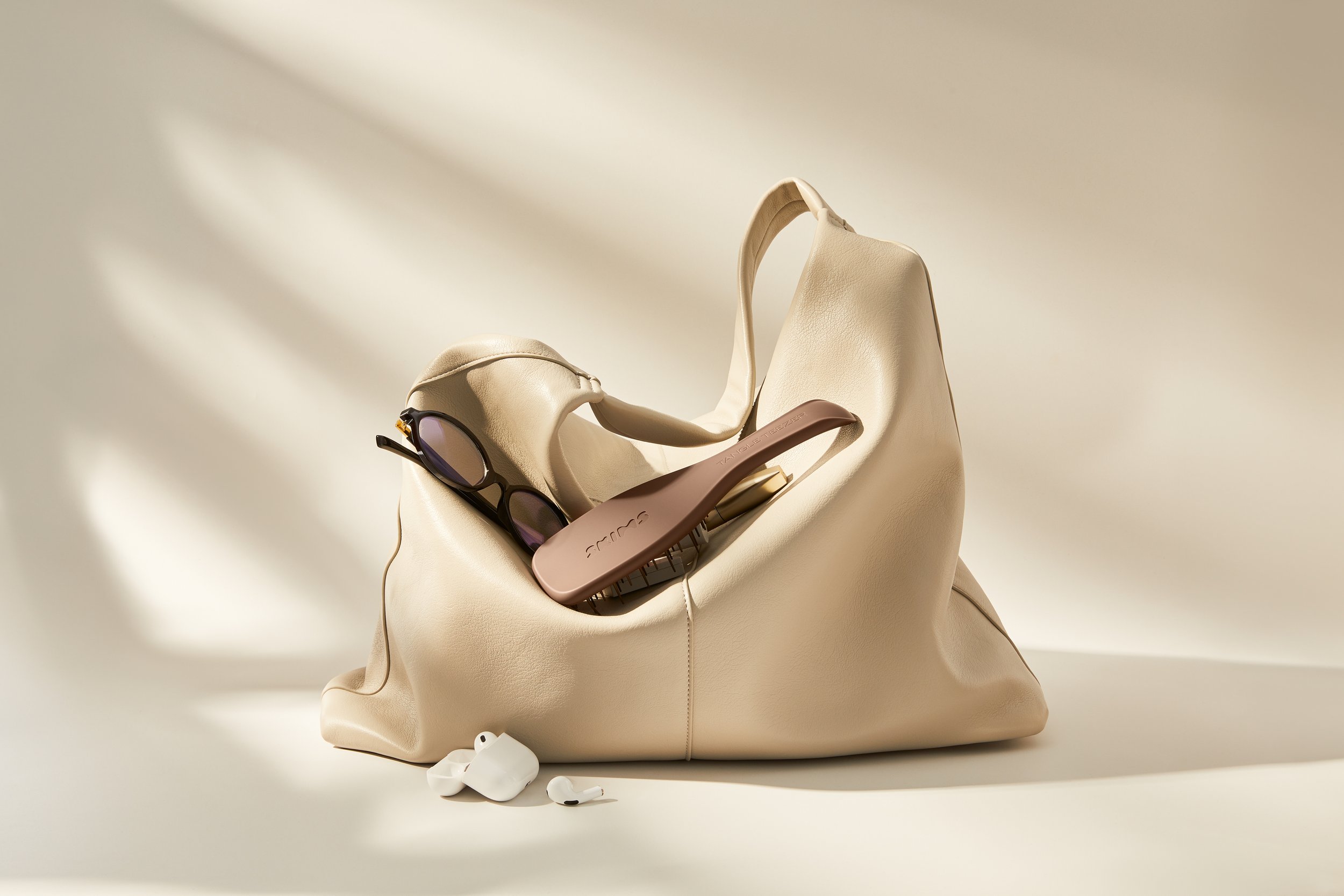 A beige slouch handbag containing reading glasses, a brown hairbrush, and white wireless earbuds, all placed on a cream surface with shadow lighting.