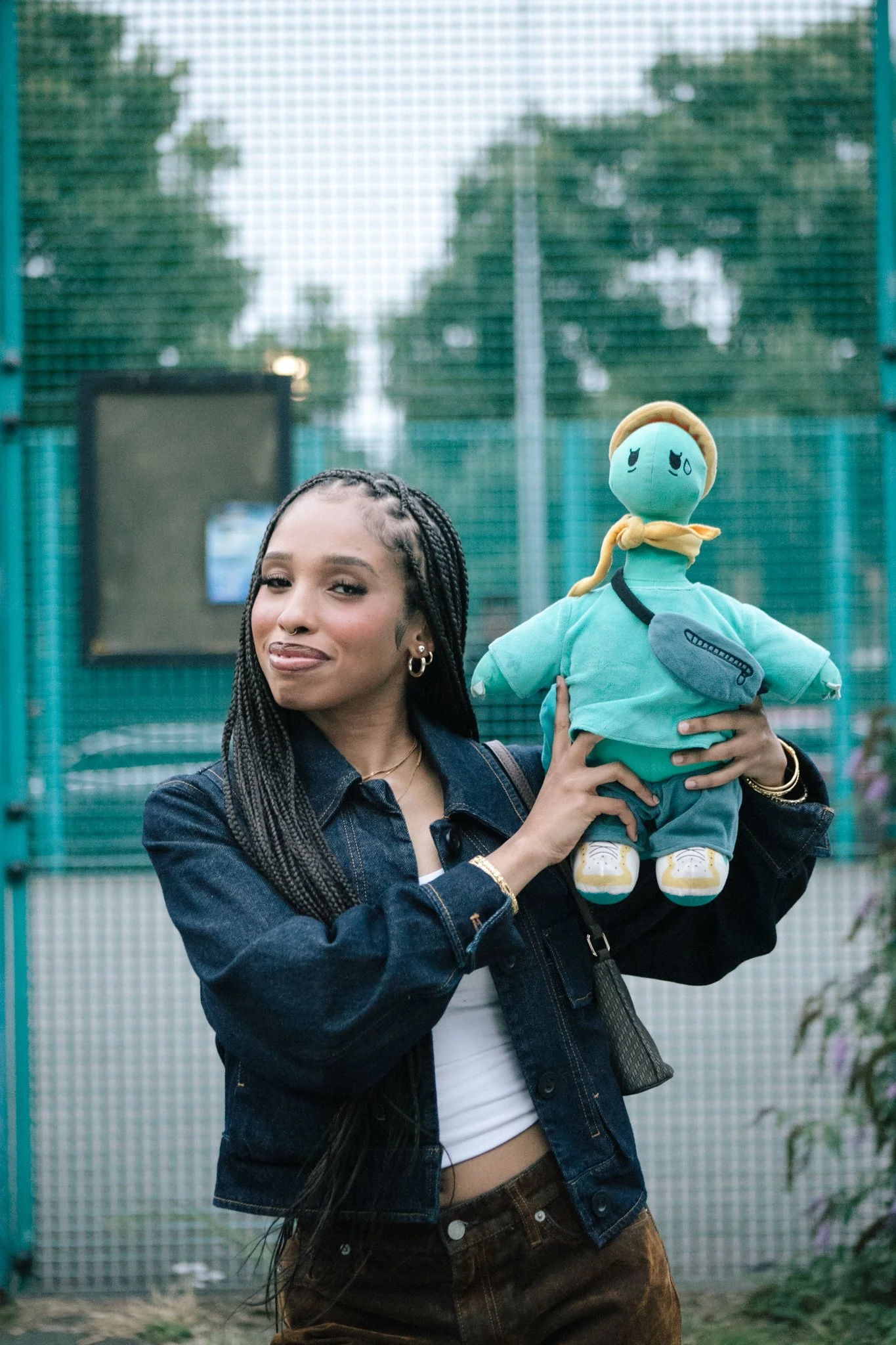 A woman with long braids holding a teal stuffed turtle plushie, that's wearing a yellow hat, outdoors against a green fence with trees in the background.