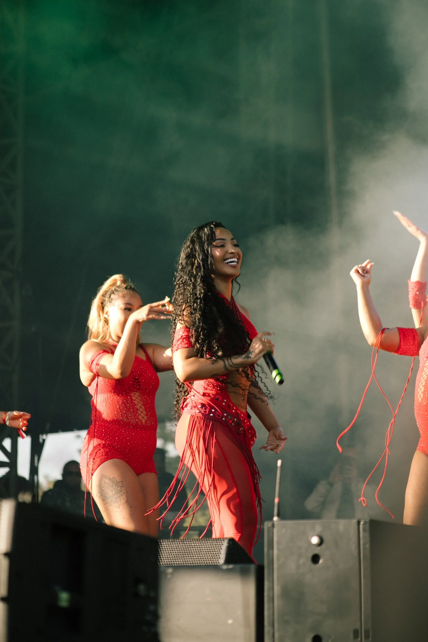 Three women dancing and singing on stage during a concert, all wearing red outfits with fringe and holding microphones. The main artist is Shenseea.